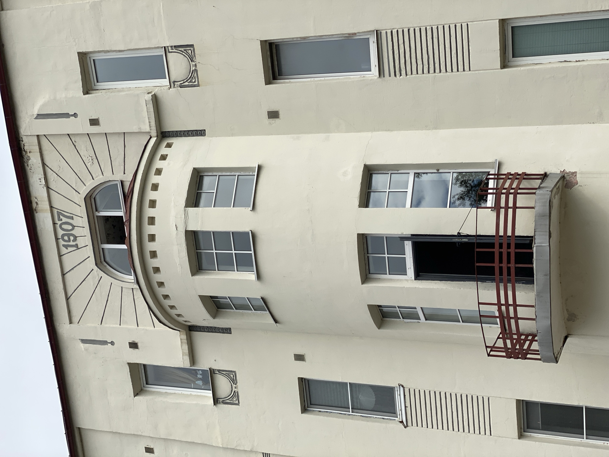 This is a medium shot, eye-level, exterior photograph of the facade of an Art Nouveau building in Sortavala, Russia. The building is painted a light cream color and features a prominent curved balcony with a red railing in the foreground. Above the balcony, a semicircular window is set into a bay that is decorated with radiating lines. The year "1907" is carved into the wall above this window. Numerous rectangular windows of varying sizes are visible across the facade. Some windows are dark, suggesting they are closed or viewed from the exterior, while others reflect the overcast sky or interior details. The building exhibits architectural detailing such as decorative ledges and vertical ribbed panels. The overall impression is of a historic residential or administrative building.