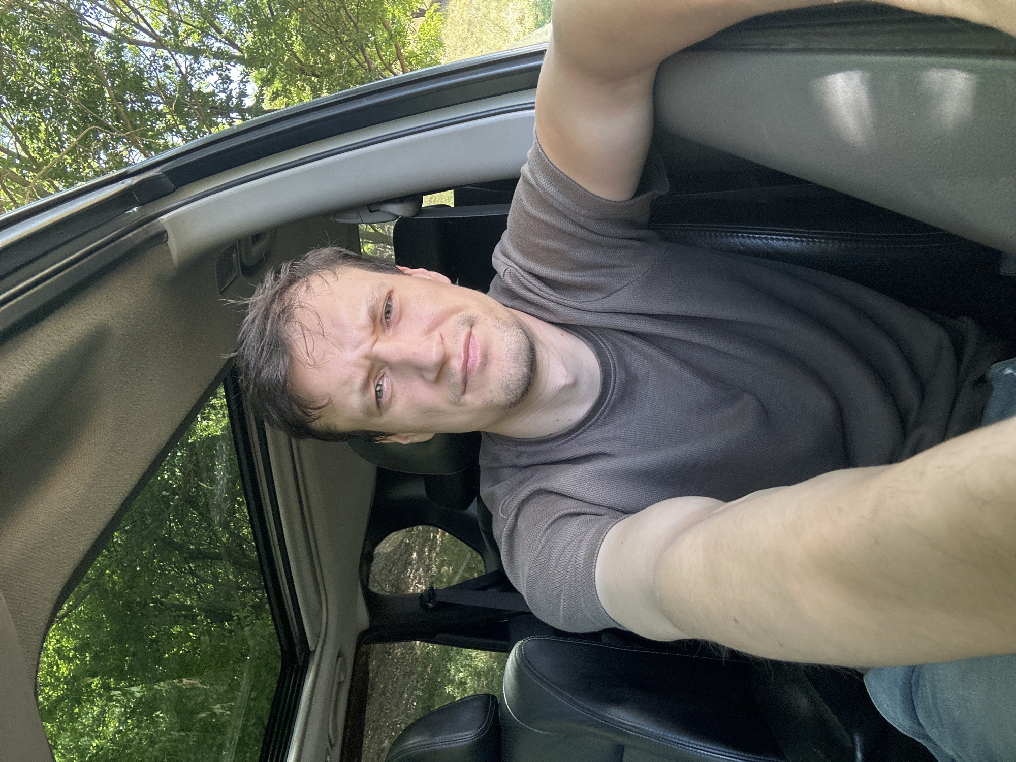 A man is taking a selfie from the driver's seat of a car, with a sunroof open and trees visible through it. The man has short brown hair, stubble, and is wearing a dark grey t-shirt and blue jeans. He is looking at the camera with a slightly furrowed brow. The interior of the car appears to be black leather. The car is parked outdoors, with greenery and trees visible outside. The image was taken in Makiv, Ukraine.