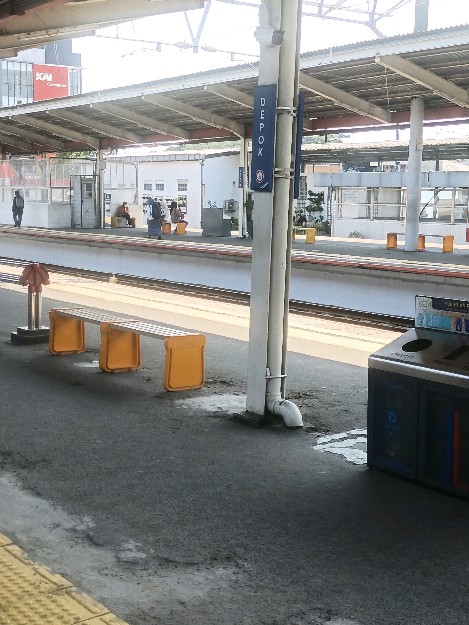 The image depicts a train station platform in Depok, Indonesia, likely the Depok railway station as indicated by a blue sign with "DEPOK" written vertically. The platform is covered by a metal roof supported by poles, and natural light streams in from above, suggesting it is daytime. In the foreground, a long, bright orange bench with a silver seat rests on the asphalt. To the right, a blue recycling bin with labels "KCI" and "Yuk Pilah P" is visible. In the background, several people are seated on orange stools and yellow benches under the covered platform, waiting. A person in a dark shirt and backpack is walking away from the camera on the left side of the platform. The environment appears clean and functional, characteristic of a public transportation hub.