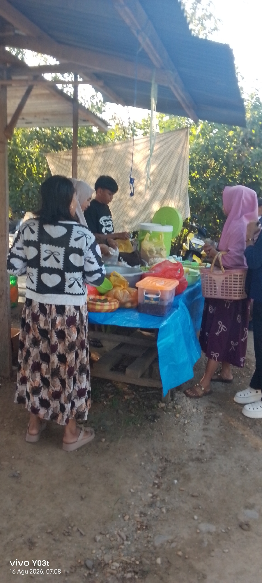 The image captures an outdoor scene at what appears to be a food stall in Palosilae, Indonesia. A table covered with a blue plastic cloth displays various food items and containers. Three individuals are interacting around the table. On the left, a person wearing a black and white patterned sweater and a long, patterned skirt stands facing away from the camera, looking at the table. In the center, a young person in a black t-shirt, with short dark hair, is engaged in preparing or serving food. To the right, a person wearing a pink hijab and a purple patterned skirt is holding a woven basket. The background consists of trees and a simple structure with a corrugated metal roof. The ground is unpaved, with small stones and dirt visible. The lighting suggests it is daytime, possibly late afternoon, with sunlight casting shadows. The overall impression is of a casual, local marketplace or roadside food vendor.