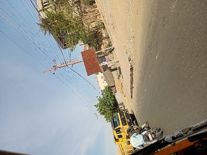 The image portrays a daylight street scene in Sharada, Nigeria, captured from a low, tilted perspective, with a partial view of an orange/yellow vehicle frame in the lower-left foreground. An asphalt road, featuring a faint white dashed line, occupies the central midground. A person wearing a light blue garment and a cap operates a black and white motorcycle, traveling away from the viewer, with a second individual in dark clothing riding as a passenger. To the left, a yellow dump truck with a black cab is visible. On the right, a concrete curb delineates the road from a dirt and gravel shoulder populated by green vegetation. In the background, a multi-story, light-colored building with dark windows stands on the right. A tall, reddish-brown utility pole, supporting numerous overhead wires, is prominently positioned on the right, adjacent to a large, rectangular, reddish-brown sign board mounted on posts, facing away from the camera. The sky is clear blue.