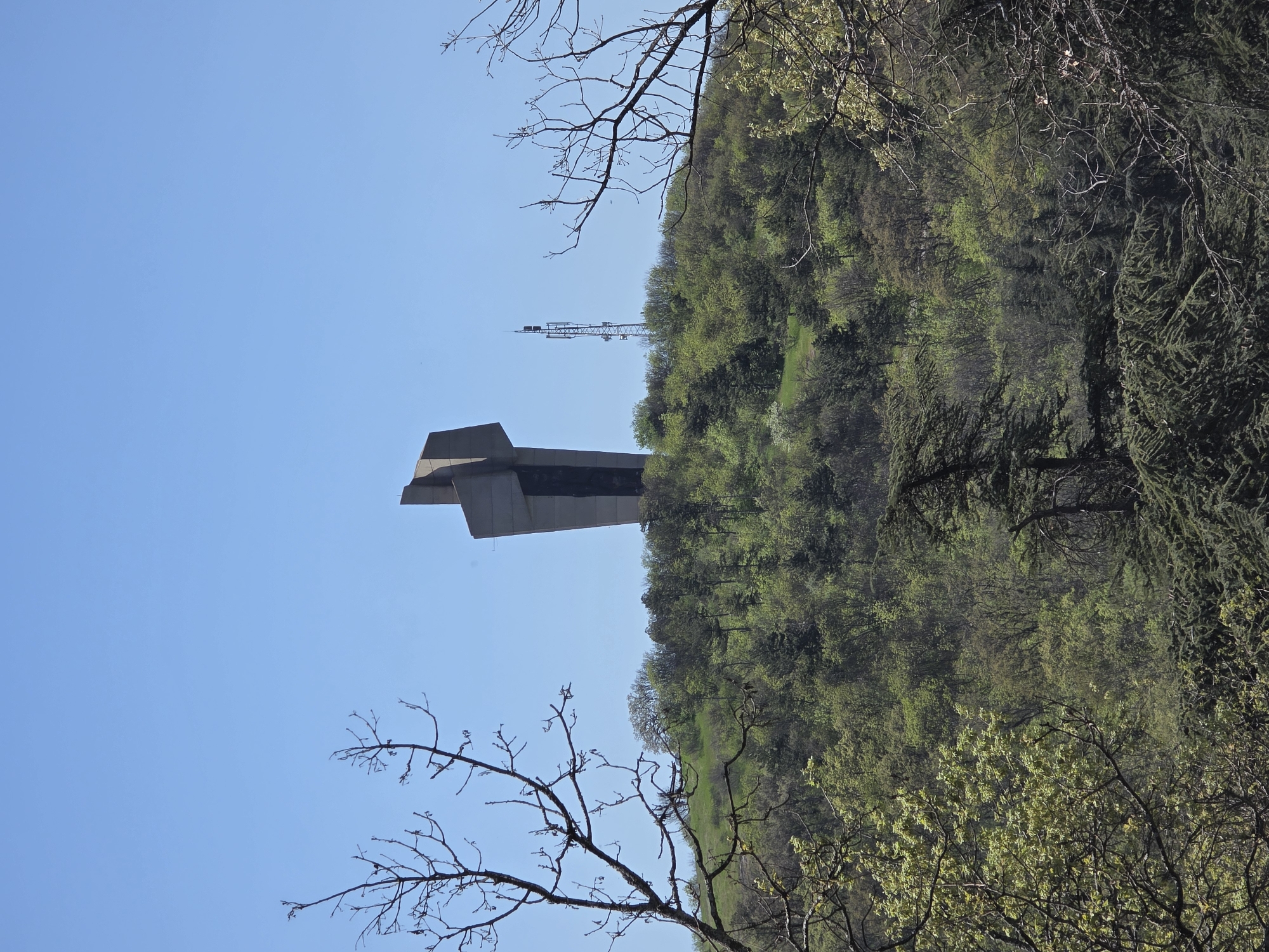 The image depicts a concrete monument in Pobeda, Bulgaria, against a blue sky. The monument has a geometric shape and is positioned near a hilltop covered with green trees. In the background, a transmission tower is visible. Leafless tree branches are prominent in the foreground, framing the monument and the landscape. The image is oriented vertically.