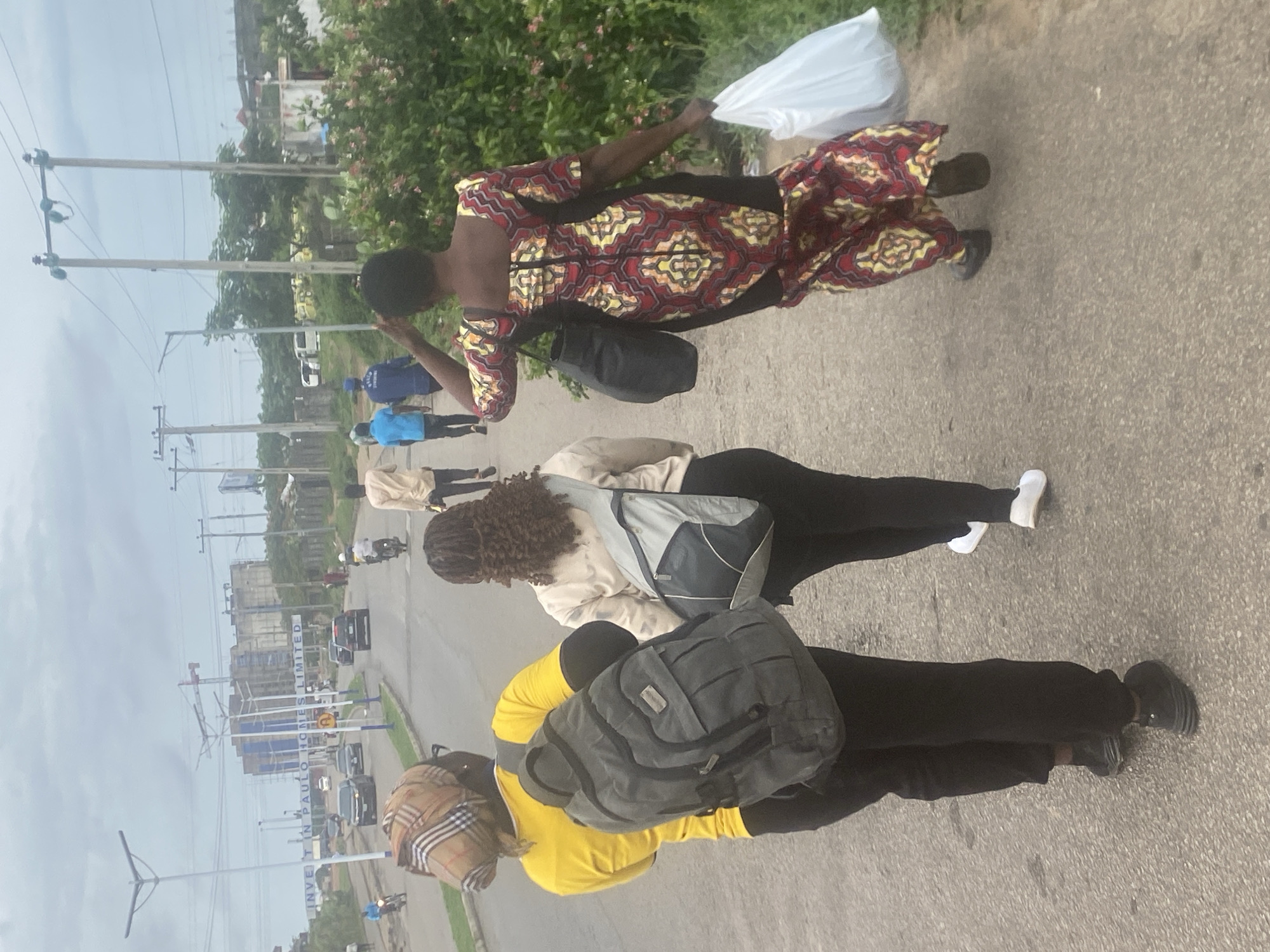 The image, taken in Lugbe, Nigeria, captures a daytime scene along a roadside under an overcast sky. In the foreground, three individuals are walking. The nearest person wears a yellow long-sleeved top, black trousers, a plaid head covering, and carries a dark grey backpack. Behind them, another person wears a light-colored jacket, black trousers, white sneakers, and has dark, curly hair, carrying a grey and black backpack. Further ahead, a person in a colorful, patterned dress (red, yellow, and black geometric design) carries a white plastic bag in one hand and a black shoulder bag.
To their left, a paved road stretches into the distance, with several cars and at least one motorcycle visible. Other pedestrians are also seen further down the road, some dressed in blue uniforms. Utility poles with power lines line the left side of the road. On the right, green vegetation, including flowering bushes, borders the path. In the mid-distance, construction activity is visible, with scaffolding and partially built structures. A blue and white sign by the road is partially legible, displaying "INVE IN PAULO HOMES LIMITED". The overall impression is of a busy urban or suburban street with ongoing development. No specific emotions are discernible from the individuals.