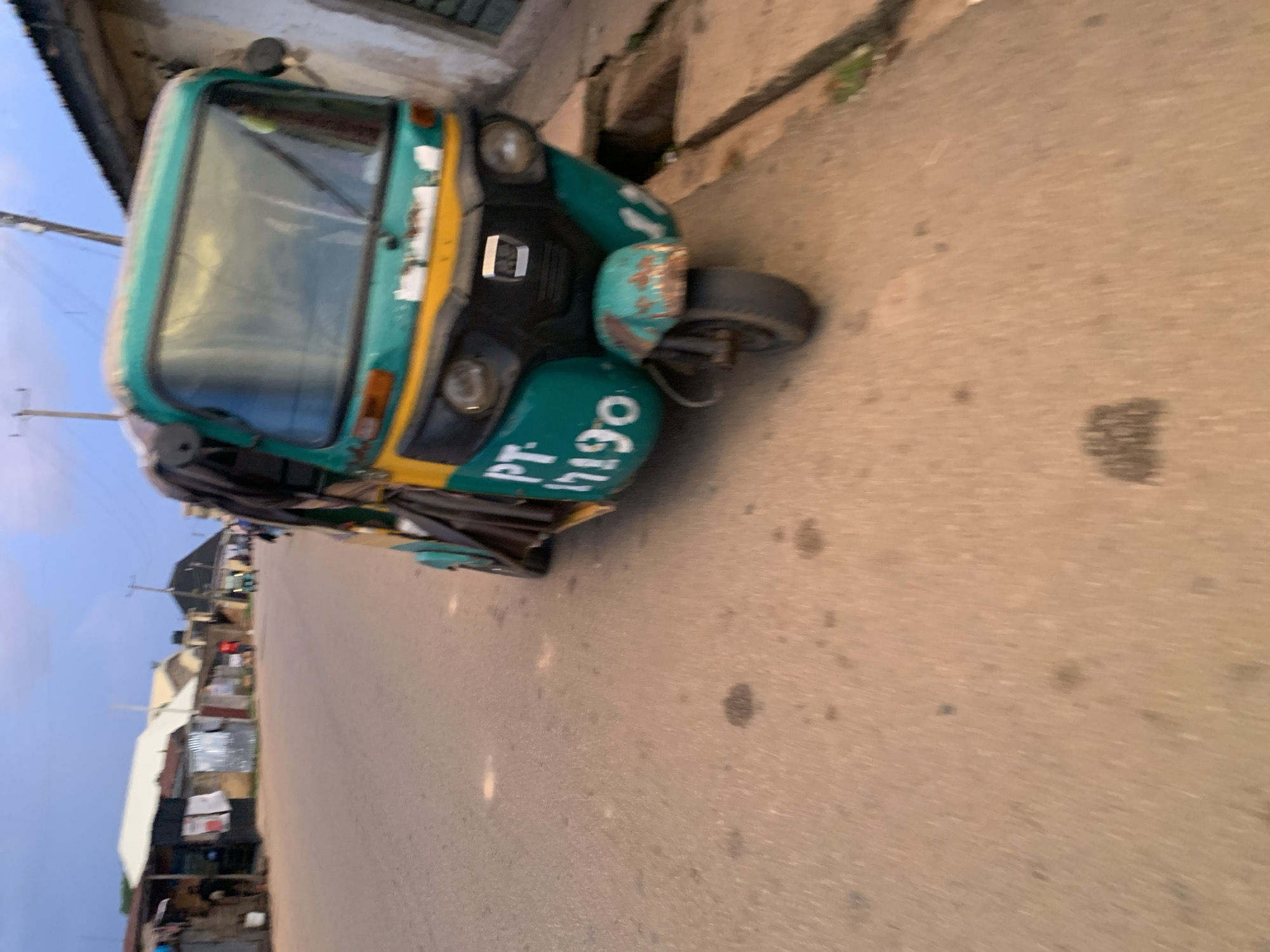 The image, rotated 90 degrees clockwise, depicts a street scene in Jos, Nigeria, featuring a prominent three-wheeled auto-rickshaw. The vehicle, commonly known as a Keke Napep, is painted green and yellow and is positioned on the left side of a paved road. Its front windshield, headlights, and front wheel are visible. On the lower front body, the registration number "PT-17190" is clearly displayed in white lettering against the green paint.
The road surface appears to be asphalt or tarmac, showing some dark stains. Along the left side of the road, a line of modest buildings with various architectural styles, including some with corrugated iron roofs, can be seen, indicating a residential or commercial district. Utility poles and power lines extend into the light, overcast sky, which suggests either an early morning, late afternoon, or a generally cloudy day. There are no people visible, and no discernible activity is taking place beyond the stationary vehicle.