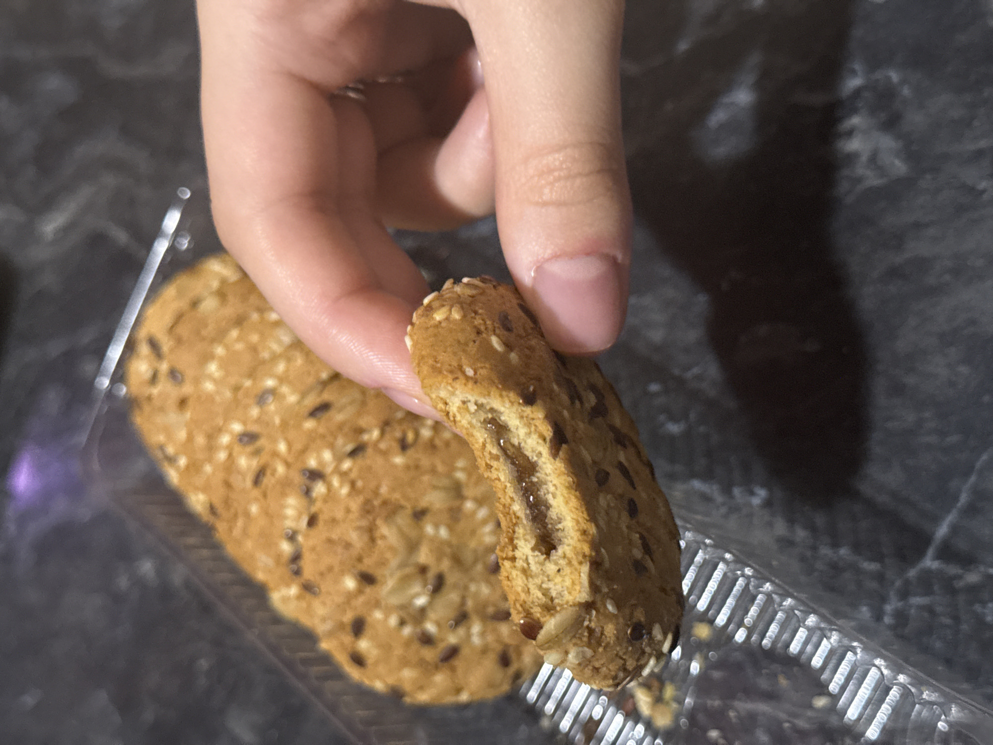 A close-up, slightly overhead shot shows a hand holding a broken biscuit filled with a dark jam, revealing the texture of the biscuit and its contents. In the background, a row of intact biscuits, also filled with jam and sprinkled with seeds, is visible within a clear plastic container. The biscuits are arranged in a tray, which sits on a dark, mottled surface that appears to be a countertop. The lighting casts a soft shadow from the hand and biscuits onto the surface. There are no discernable elements indicating the location in Lobacheve, Ukraine.