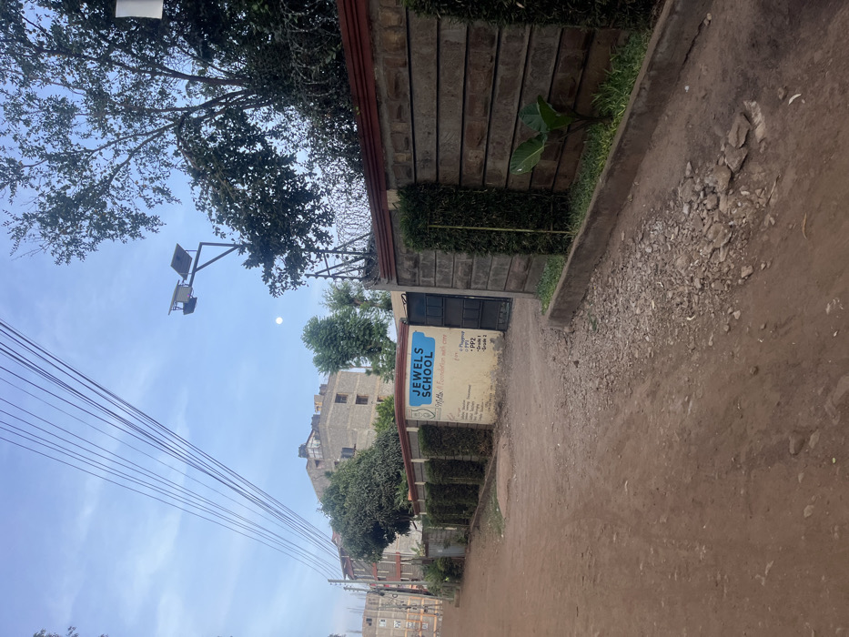This image, captured in Gitothua ward, Kenya, presents a static outdoor scene, likely during early morning or late afternoon, characterized by a bright, clear blue sky and a visible moon. The foreground is dominated by a dirt road, uneven and scattered with gravel and small rocks, particularly along its right side. A concrete curb separates the road from a neatly trimmed green hedge, positioned in front of a stone-patterned wall. This wall, topped with what appears to be barbed or razor wire, suggests security.
An entrance in the wall features a dark-colored metal gate. To its left, a blue and yellow sign is attached, prominently displaying "JEWELS SCHOOL." Below this, "Mother Foundation" is partly visible, along with academic levels: "PP1," "PP2," and "Grade 1."
Overhead, a streetlight with visible solar panels extends from a pole. Multiple power lines or utility cables stretch diagonally across the upper left sky. Lush green trees are visible, some overhanging the scene, with more foliage and several multi-story buildings in the background, hinting at a residential or institutional area. The architecture of the buildings is functional, contributing to an urban-fringe atmosphere. No explicit activity or people are present, offering a tranquil view of the local environment.