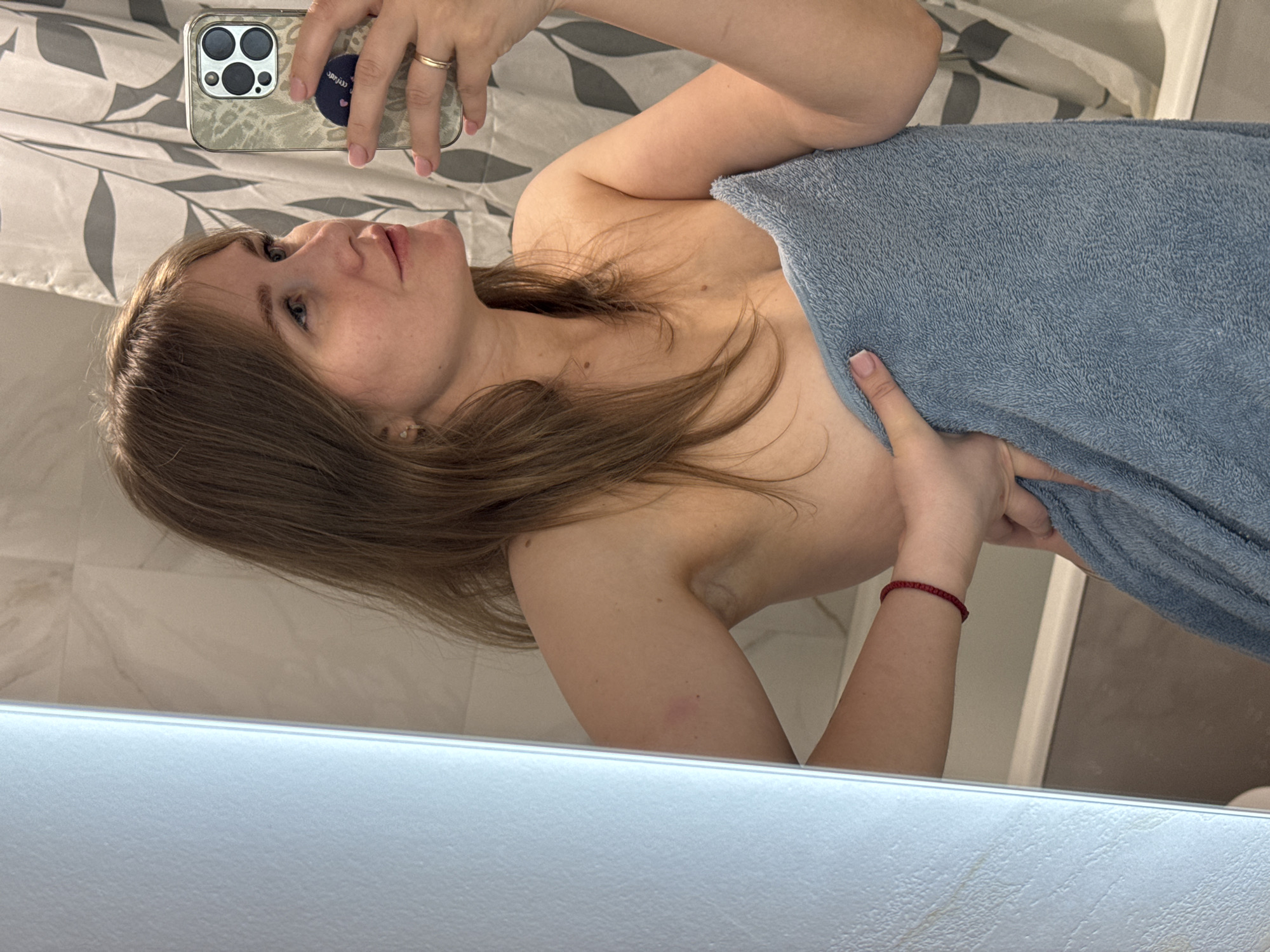 A fair-skinned woman is taking a selfie in a bathroom. She is holding a smartphone with a patterned case in her left hand, capturing her reflection in the mirror. The woman is partially covered with a light blue towel, held in place by her right hand. She has light brown hair and is wearing a gold ring. The background includes a shower curtain with a leaf pattern and tile walls. The image appears to be taken in a residential setting, possibly in Ферма, Russia, given the user's specified location.