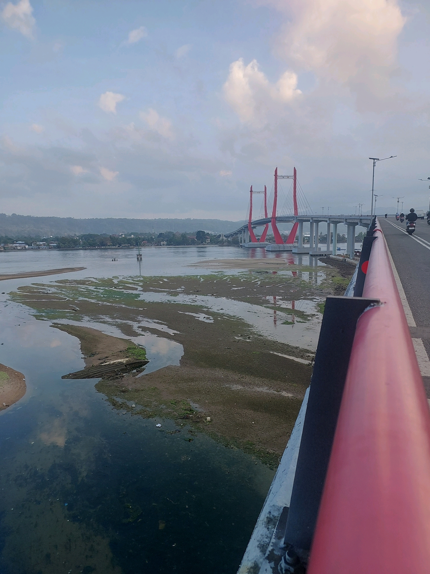 The image shows the Ambon Bay Bridge in Ambon, Indonesia, during a low tide. The bridge is a prominent cable-stayed bridge with striking red pylons. The bay's water level is low, exposing muddy shores with patches of green vegetation. A partially submerged wooden boat hull is visible in the foreground, resting on the mudflats. The water reflects the cloudy sky. The foreground features a road with a red railing, indicating the viewpoint is from the bridge. A few motorcycles and their riders are visible on the bridge, suggesting ongoing traffic. In the distance, the city of Ambon is nestled along the coastline, with hills rising behind it. The overall scene captures a typical day in Ambon, highlighting the bridge's grandeur and the natural landscape of the bay.