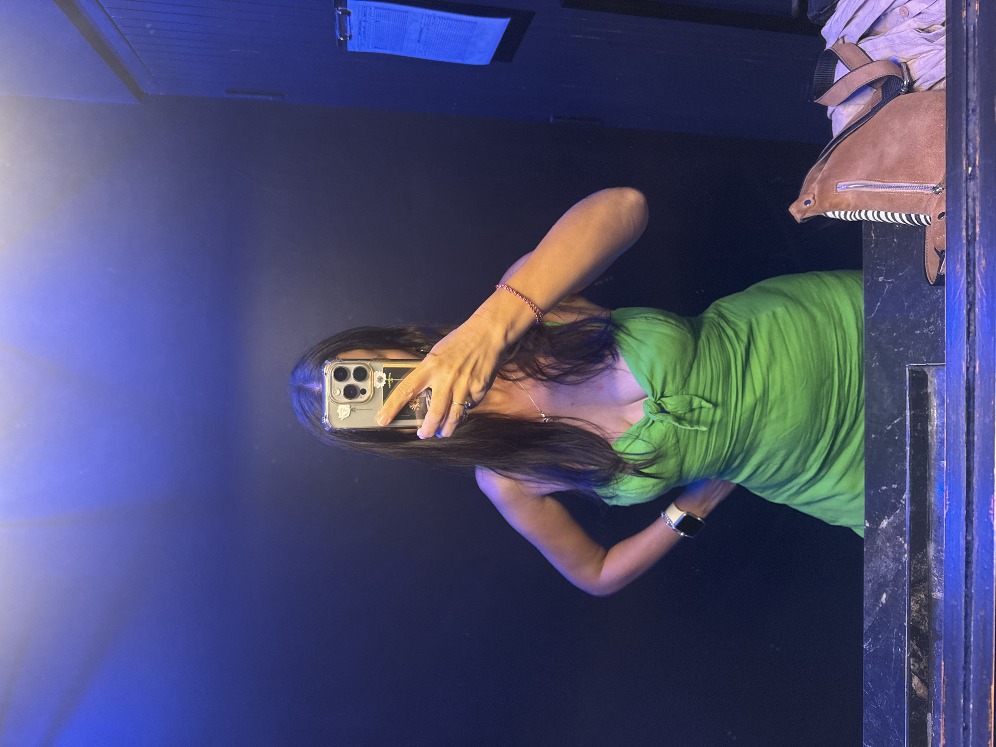 A woman stands in front of a dark blue wall, taking a mirror selfie. She is wearing a bright green strapless dress with a tie front. Her right arm is akimbo, and her left hand is holding her phone. She has long dark hair and is wearing a necklace and a bracelet. A smartwatch is visible on her left wrist. In the foreground, on a dark marbled surface, is a brown suede bag with a striped lining and a zipper. To the right of the woman, a blue clipboard with papers is attached to a wooden surface. The environment appears to be an indoor setting, possibly a bathroom or dressing room, with a blue light illuminating the upper portion of the wall. The overall scene is dimly lit, with the exception of the bright green dress and the blue light.