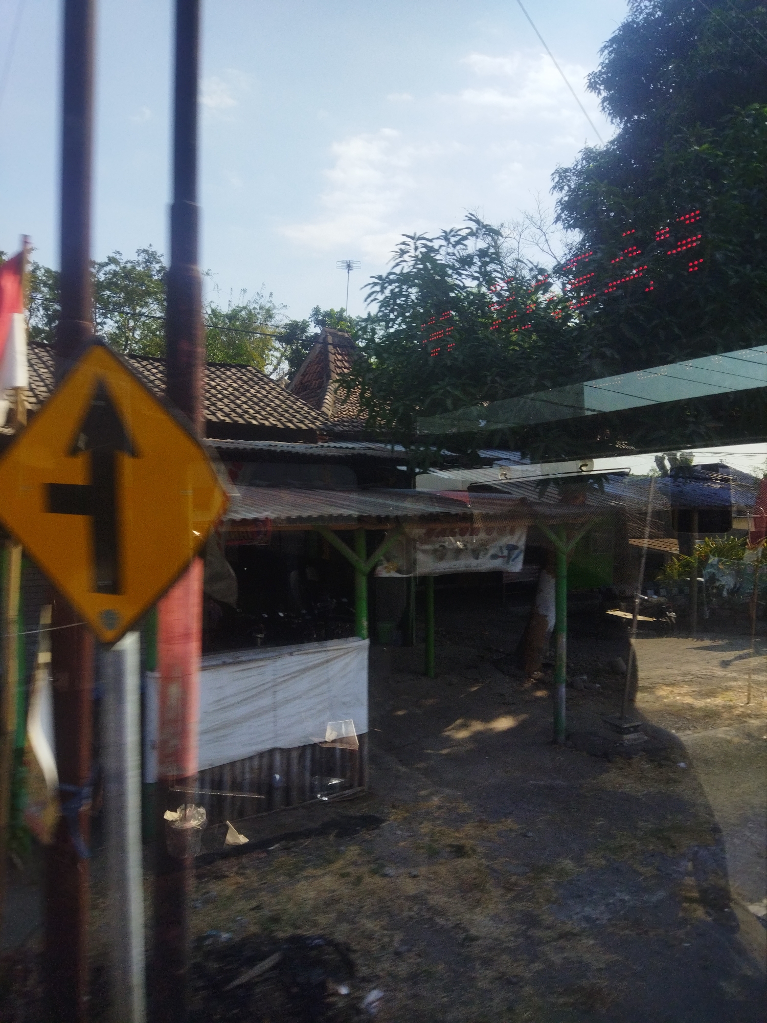 The image shows a street scene in Nguter, Indonesia. In the foreground, a yellow diamond-shaped traffic sign with a black arrow indicating a turn to the left and straight ahead is visible, mounted on a pole. Behind the sign, a row of small roadside structures with corrugated metal roofs can be seen. Some of these structures appear to be shops or stalls. Trees with red, berry-like decorations are present in the background, with a clear blue sky above. The ground is dirt and sparsely covered with dry grass. The scene appears to be along a road, with a paved surface visible in the mid-ground and background. The overall impression is of a typical rural or semi-rural environment in Indonesia.