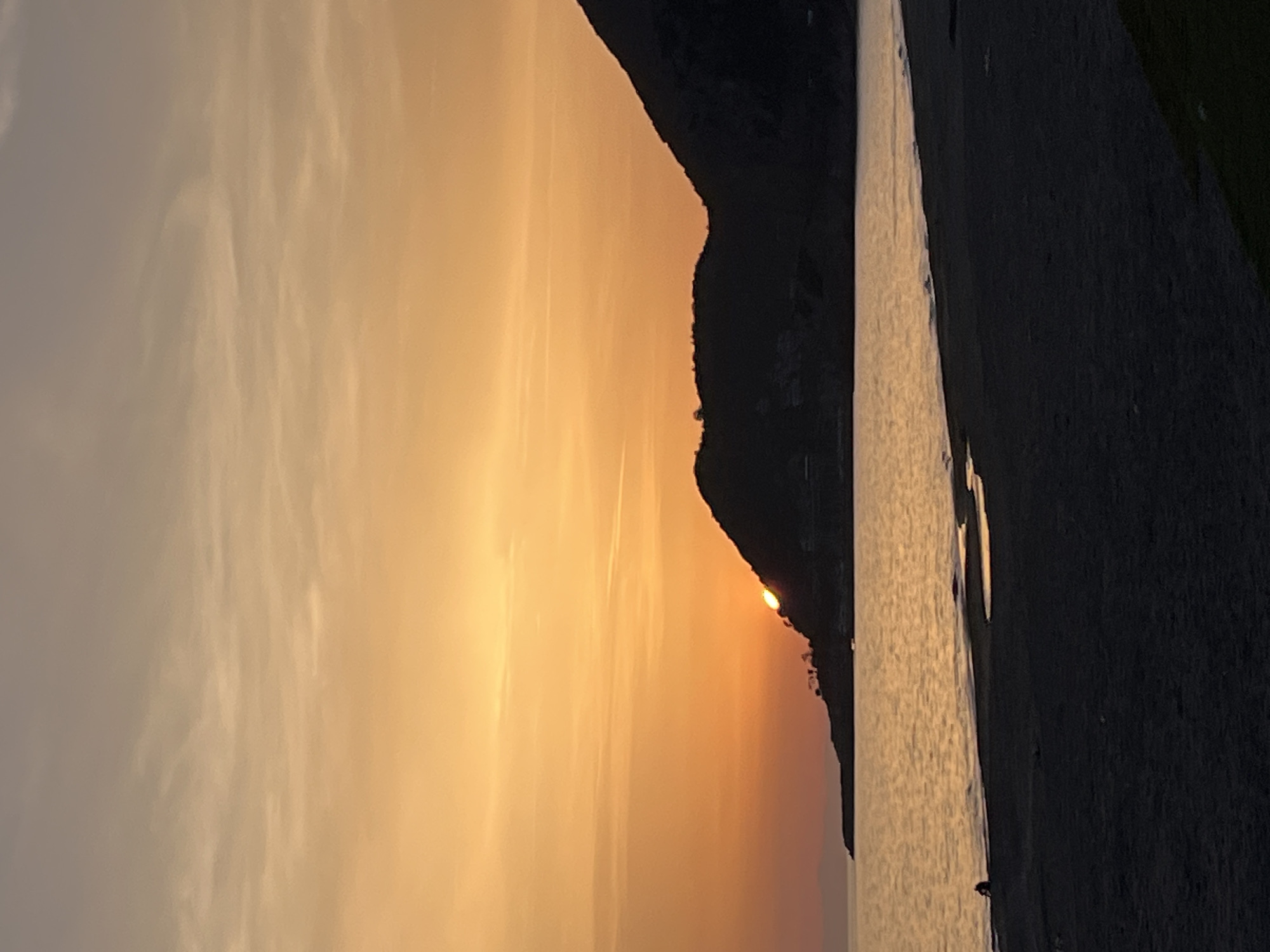 The image depicts a sunset at a beach, likely in Alanya, Turkey. The sun is setting behind a dark, mountainous headland. The sky transitions from light to dark, with hues of orange and yellow. The sea reflects the sunset colors. A beach is on the right side of the image. There is a vague outline of a person on the beach.