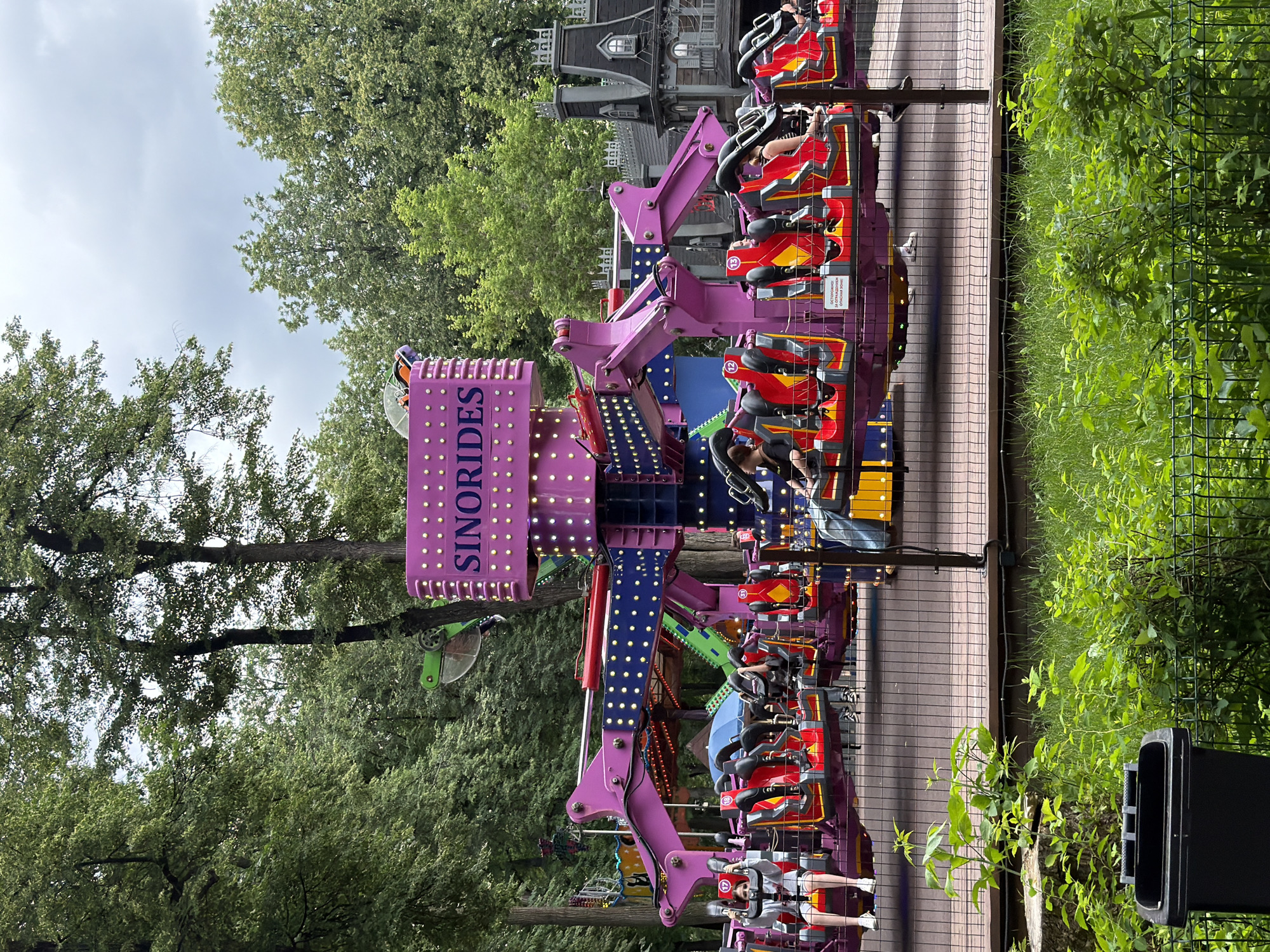 The image shows a colorful amusement park ride with people seated in it. The ride is a large, mechanical structure with purple and red accents and numerous lights. Some people are visible in the seats, appearing to be enjoying the ride. The setting appears to be an outdoor amusement park with trees and a cloudy sky visible in the background. The ride has a sign that says "SINORIDES" in large, purple lettering. There is also some text on a warning sign on the side of the ride, but it is too small to be legible. The overall atmosphere seems lively and fun, characteristic of an amusement park. The time of day appears to be daytime, with overcast weather conditions.