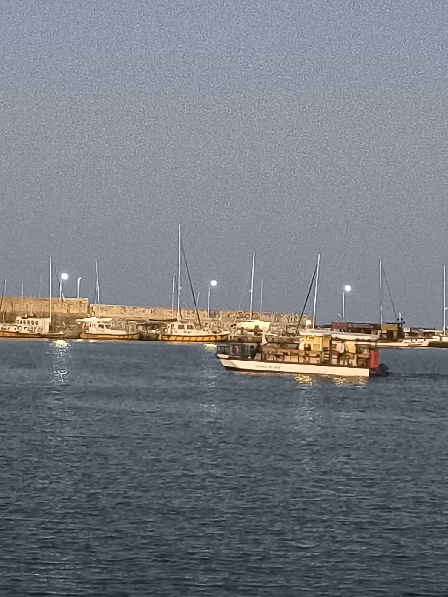 The image depicts a harbor scene in Varna, Bulgaria, with several boats docked near a stone pier. In the foreground, the dark blue water reflects the lights from the boats and the pier. The main subject is a large, elongated boat with a tan and white superstructure, heavily laden with what appear to be goods or supplies, possibly for transport or sale. This boat is positioned towards the right side of the frame, closer to the viewer. To the left, a line of other boats, mostly smaller and some with masts, are moored along the pier, which is constructed from weathered, light-colored stones. Some of these boats also have tan and white exteriors. The sky above is a clear, muted gray-blue, indicating either early morning or late evening light. There are no people clearly visible in the image. The overall impression is one of a working harbor, with boats engaged in or ready for their daily activities.