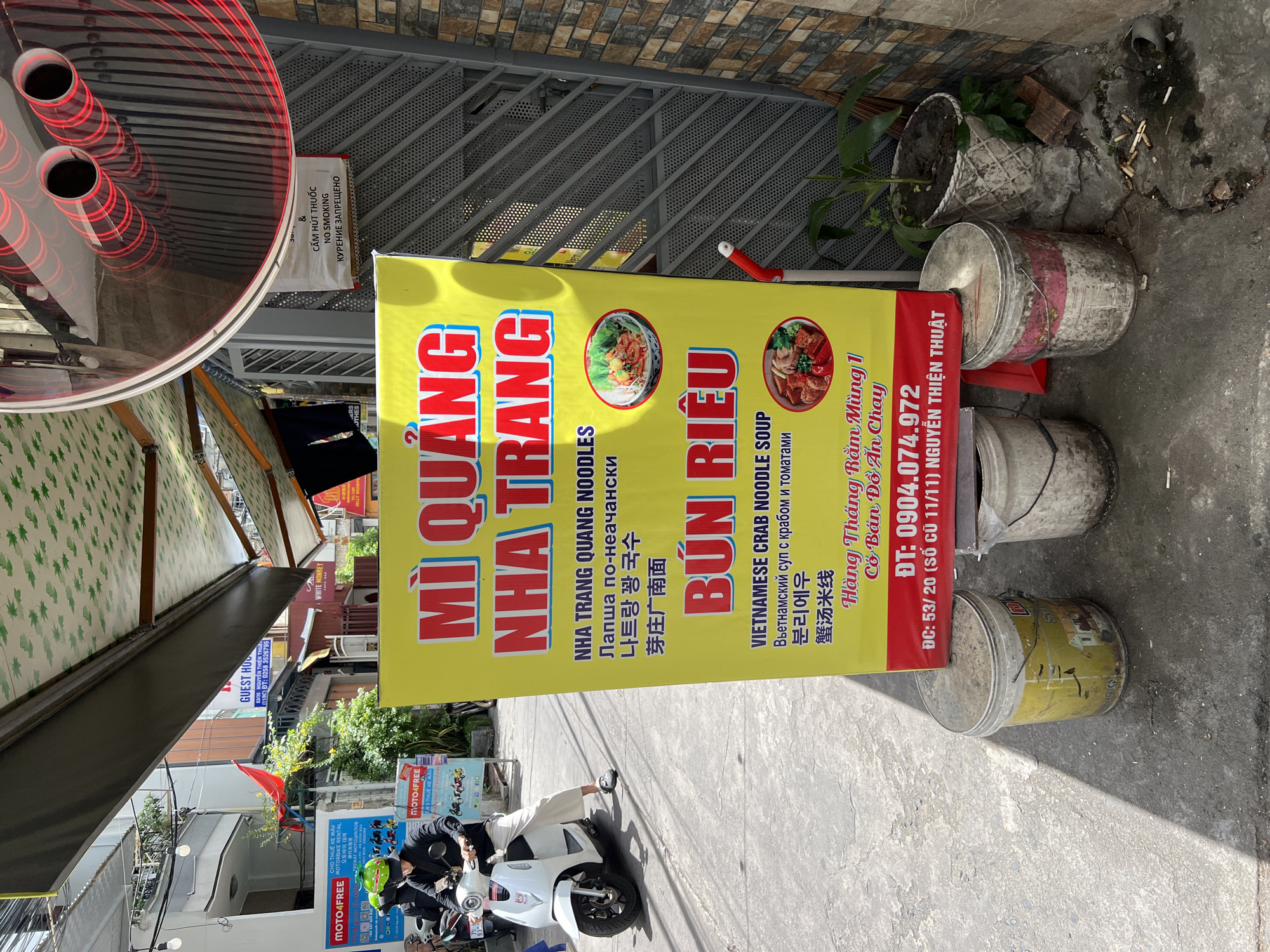 A street scene in Nha Trang, Vietnam, shows a vibrant yellow sign advertising "Mì Quảng Nha Trang" and "Bún Riêu" with accompanying images of noodle dishes. The sign also displays text in Vietnamese, Russian, Korean, and Chinese, along with a phone number and address. To the left of the sign, a person wearing a green helmet rides a white scooter. In the background, buildings line the street, and awnings extend over shopfronts. To the right of the sign, a "No Smoking" sign is visible. Several discarded buckets and cigarette butts are present on the ground, suggesting a public outdoor setting. The lighting indicates it is daytime.