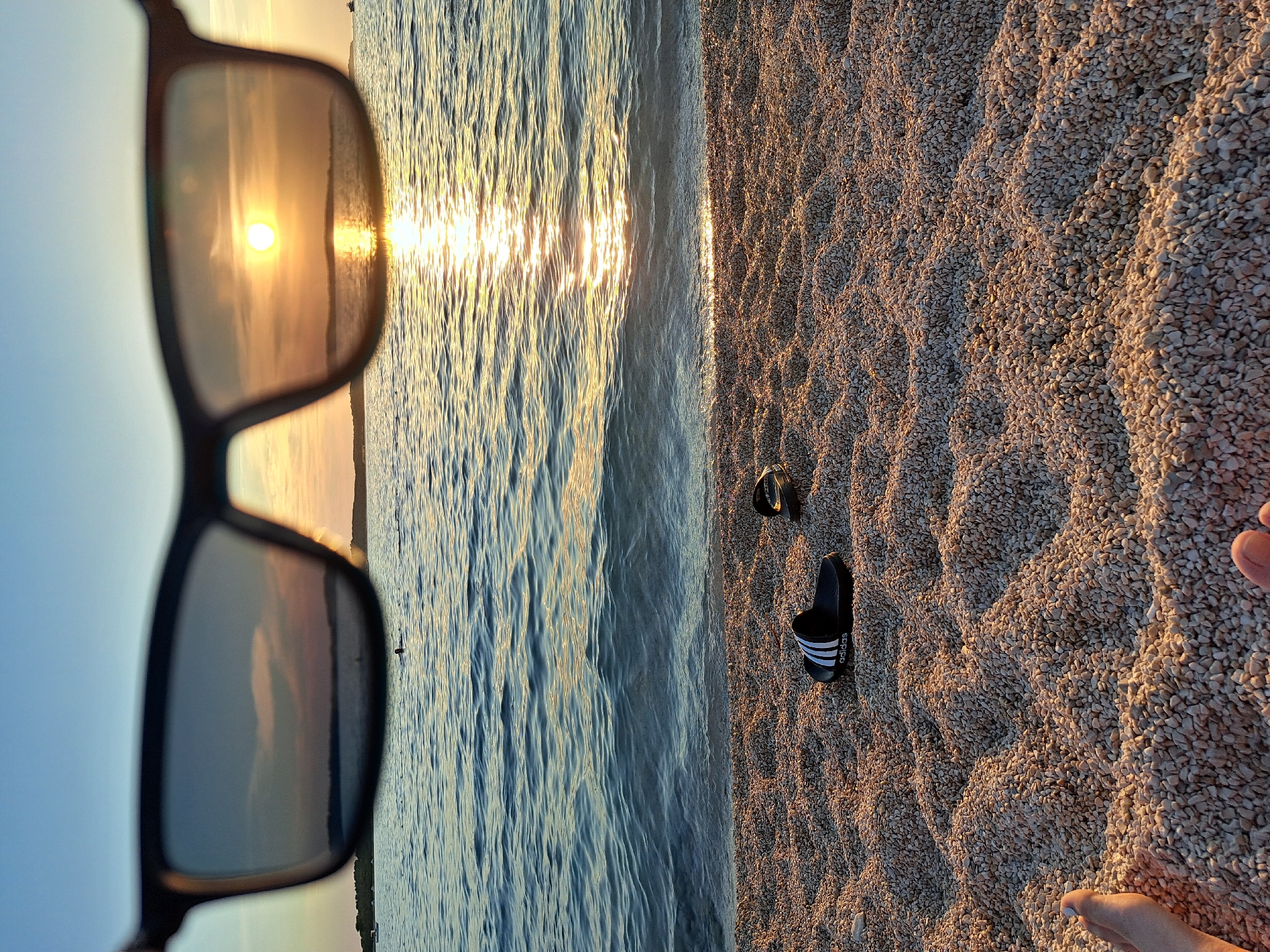 This media file depicts a beach scene at sunset. The camera is positioned low, looking across the shoreline. In the foreground, a pair of sunglasses is held up, reflecting the setting sun and the sky. The sun appears as a bright, warm orb with rays of light shimmering on the water's surface.
The scene transitions to the water's edge, where gentle waves lap against a pebbly beach. The pebbles are light in color, with some darker stones interspersed, creating a textured surface. Scattered on the pebbles are two pairs of black Adidas slide sandals with white stripes. Further along the beach, the feet of at least two individuals are visible, suggesting they are relaxing by the sea. One set of feet shows a person with toenails painted white.
The overall atmosphere is one of tranquility and relaxation, typical of a sunset by the sea. The warm, golden light of the setting sun bathes the scene, creating a serene and inviting ambiance. The sky is a pale blue, with hints of pink and orange reflected in the sunglasses and on the water. The presence of the sunglasses and sandals suggests a casual outing, likely a vacation or a leisurely evening stroll. There is no discernible text in the image, apart from the "adidas" logo on the sandals.