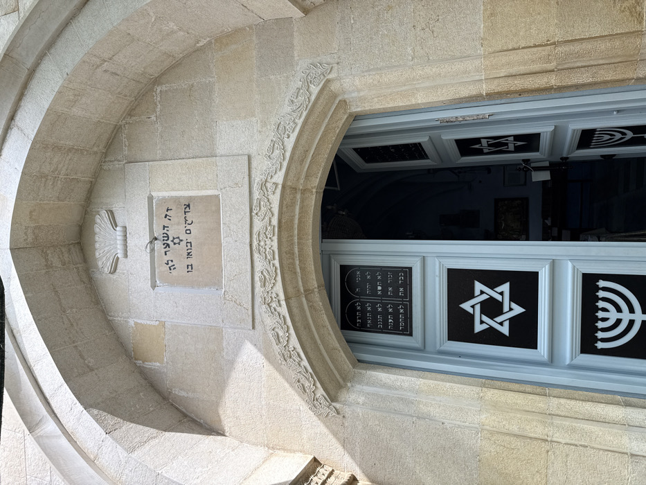 The image displays the arched entrance of a building in Safed, Israel, likely a synagogue given its iconography. The entrance is constructed from light-colored, cut stone blocks. Above the double doors, a rectangular stone plaque is set into the wall, featuring Hebrew script and a central Star of David symbol. A carved shell motif is positioned directly above this plaque. An ornamental stone frieze with a continuous leafy pattern frames the arch directly above the door frame.
The double doors are painted a light blue or pale grey. Each door panel contains three distinct framed sections. The uppermost section on both doors displays a rectangular panel with Hebrew text, including "אנכי ה'". The middle section on each door features a white Star of David on a dark background. The lowermost section on each door presents a white seven-branched Menorah symbol, also on a dark background. The right door is partially ajar, revealing a glimpse into a dimly lit interior with a dark blue vaulted ceiling and a framed object or artwork visible on a distant wall. The exterior stone is brightly illuminated by daylight. No individuals are present in the scene.