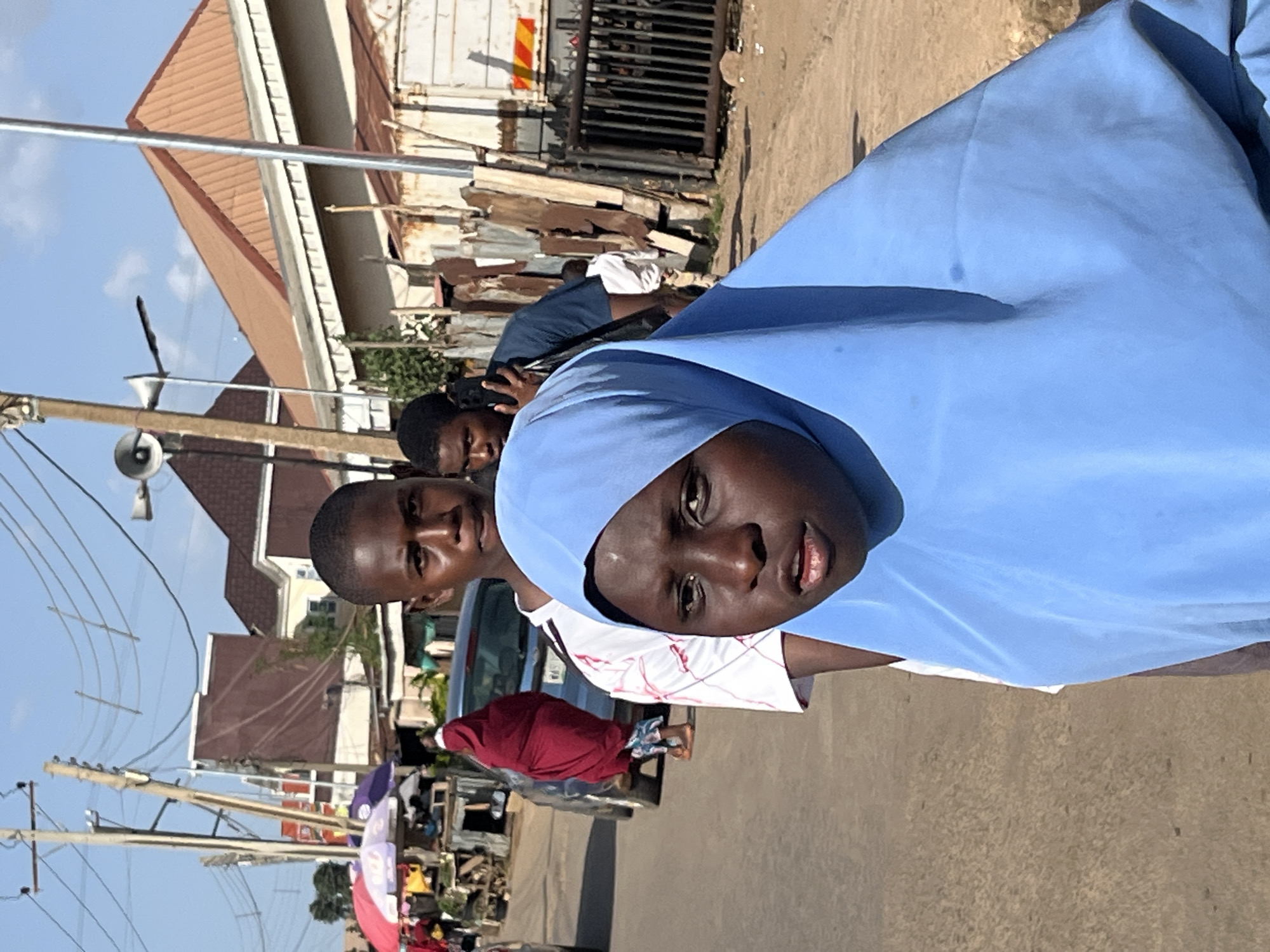 The image shows a street scene in Jos, Nigeria, under a clear blue sky. In the foreground, a young person wearing a bright blue hijab is the primary subject, with their face turned towards the camera. Behind them, a young male is visible, looking forward, and another person is partially visible talking on a mobile phone. In the background, there is a busy street with parked cars, market stalls with umbrellas, and various buildings. The environment appears to be an urban setting with a mix of residential and commercial structures. The ground is unpaved and dusty. Power lines and utility poles are prominent in the upper portion of the image, indicating urban infrastructure.
