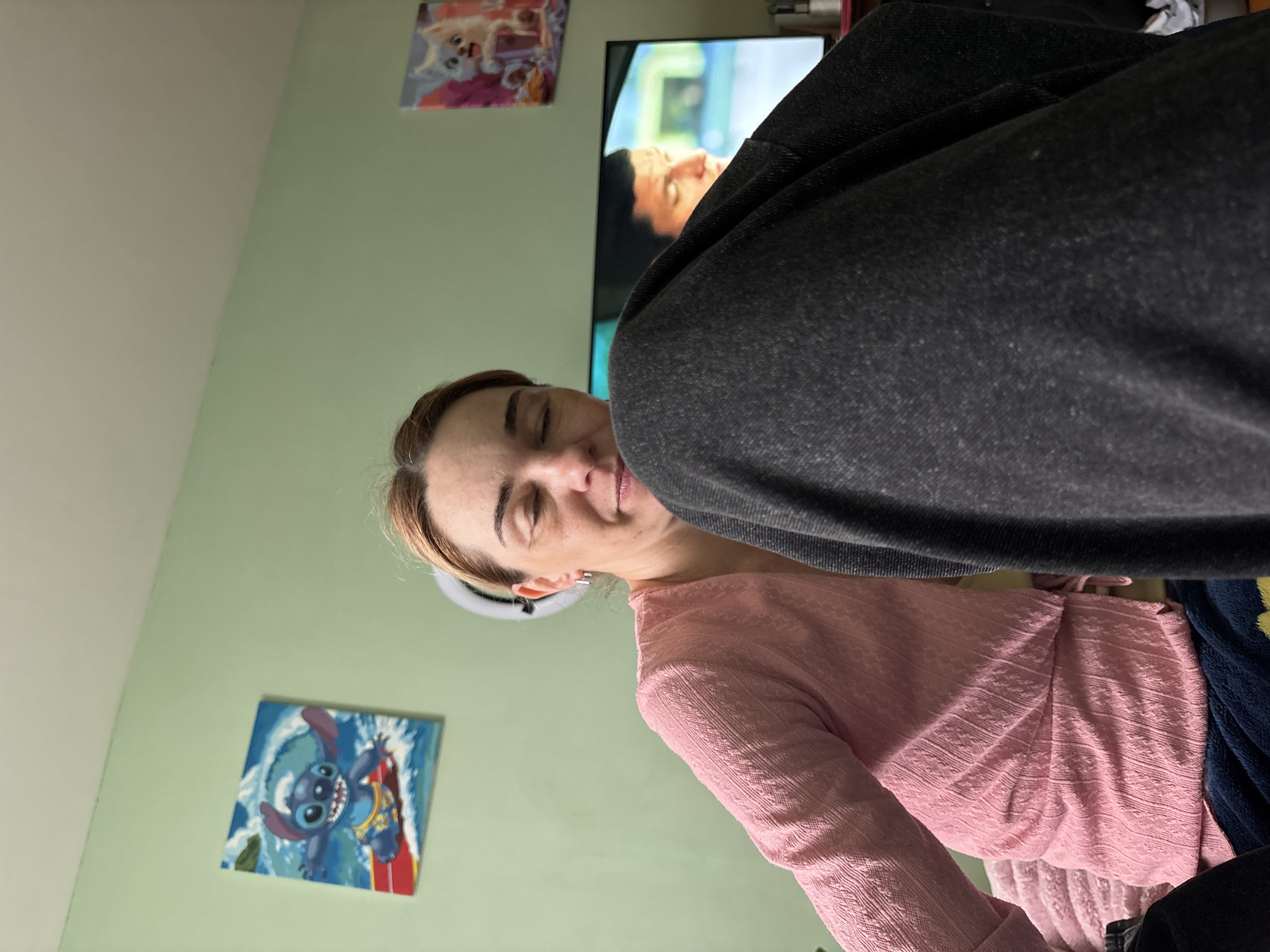 A woman with light brown hair and a pink shirt is in a room with light green walls. There is a TV behind her showing a man's face. There is a dark grey object in the foreground, obscuring part of the scene. On the wall are two pictures, one of Stitch and another of a dog. There is a ring light next to the woman's face. The location is possibly Murino, Russia.