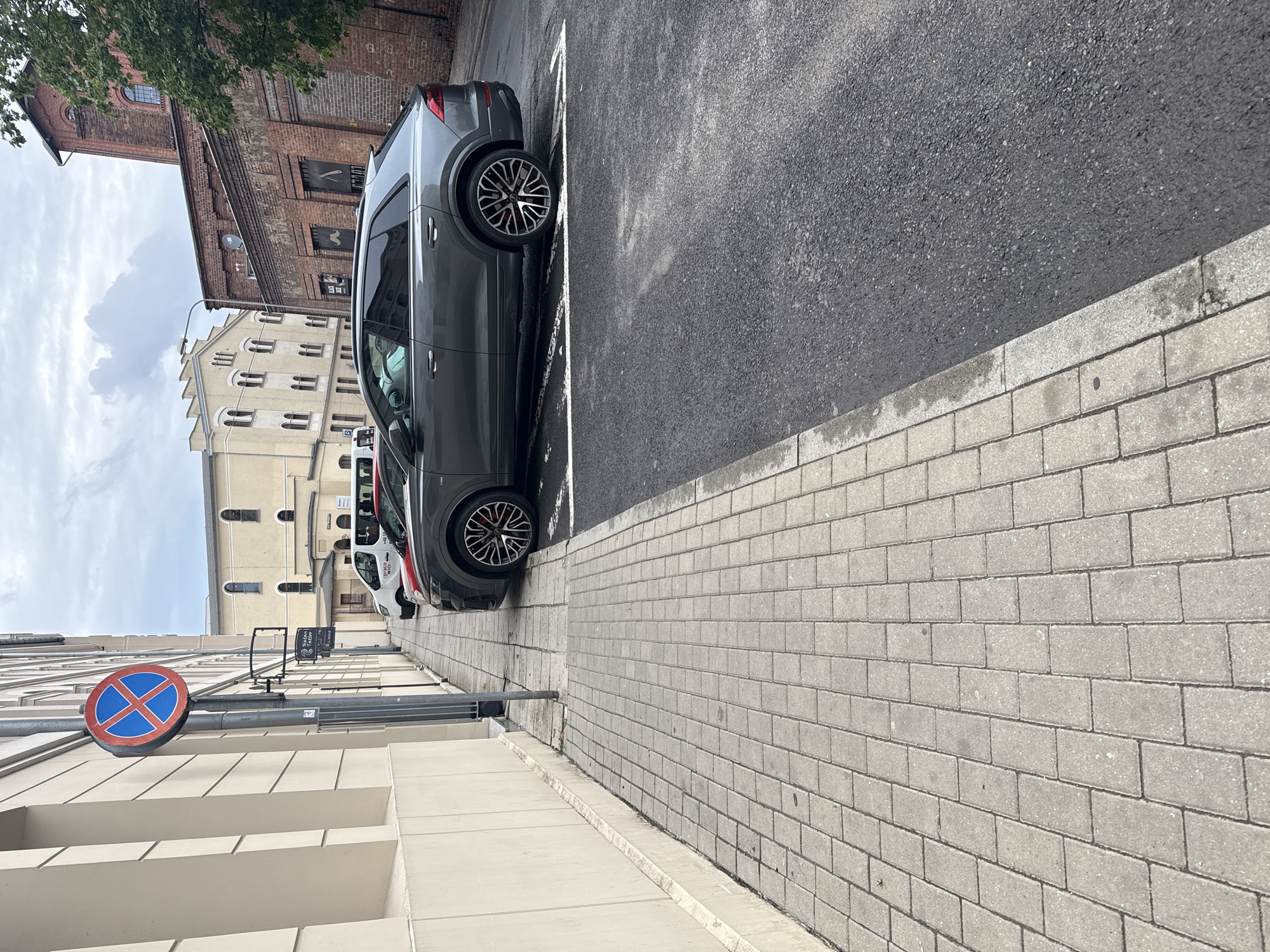 The image shows a street scene in Zielona Góra, Poland. The perspective is from a low angle, looking up towards a gray SUV parked on the right side of the frame, which is oriented vertically. The car is parked in a designated spot marked by white lines on asphalt. In the background, there are several buildings: a red brick industrial building on the left, a cream-colored building with arched windows in the middle ground, and a white van is visible behind the SUV. A street sign with a red circle and a blue cross inside is visible on the left, indicating a prohibition. The weather appears to be overcast, with a cloudy sky. The ground is paved with light gray bricks, and there is a concrete sidewalk. The overall scene depicts an urban environment, likely during the daytime. No people are visible in the image. The visible text on the white van is "EURO-WIEL". There is also text on a sign in the background which is difficult to read clearly.