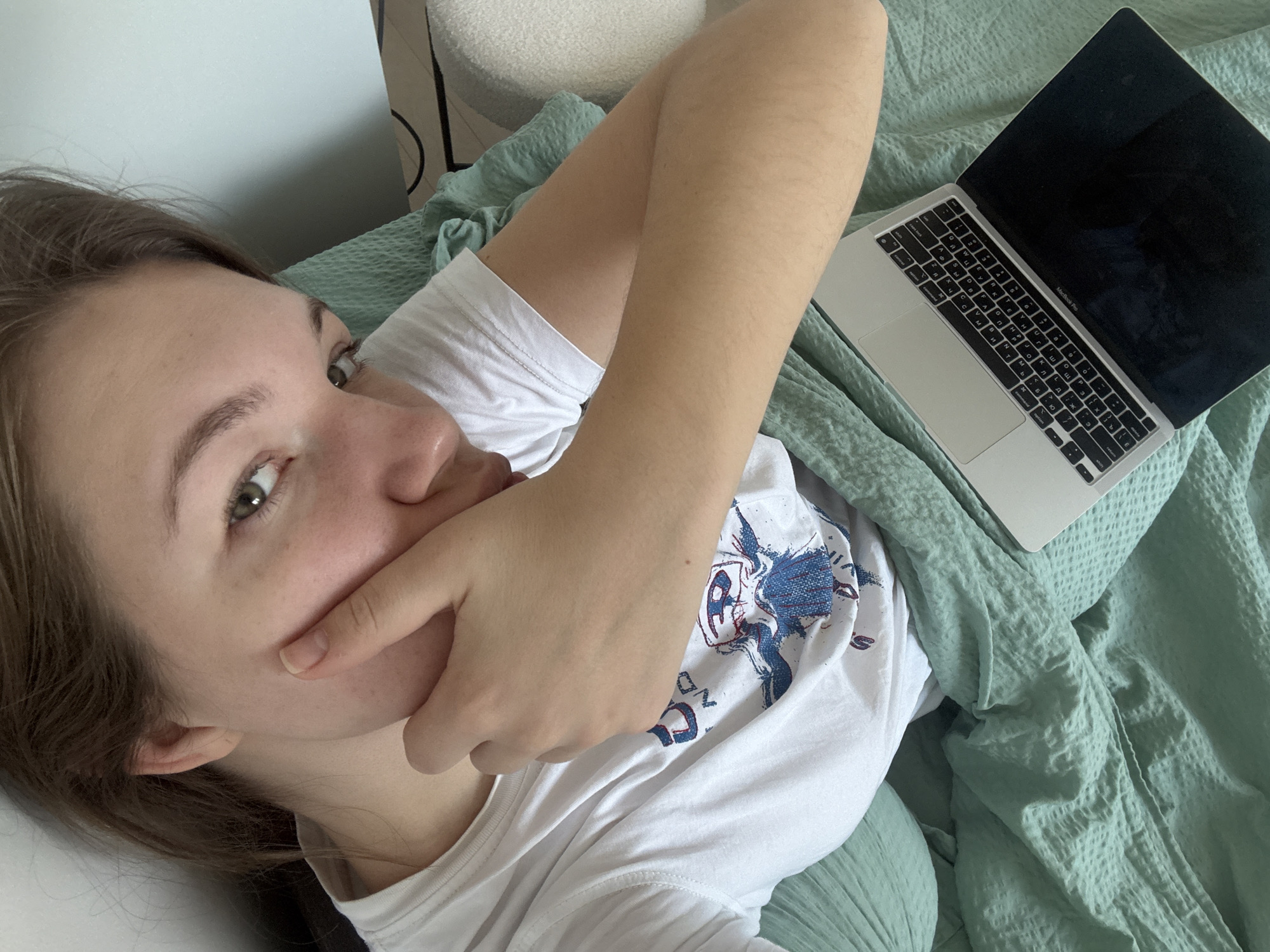 A young woman is lying on a bed with a green waffle-patterned blanket. She is wearing a white t-shirt with a blue and white graphic. Her right hand is covering her mouth. A laptop is open on the bed to her right. The background includes a white wall and a white armchair. The location cannot be determined from the image.
