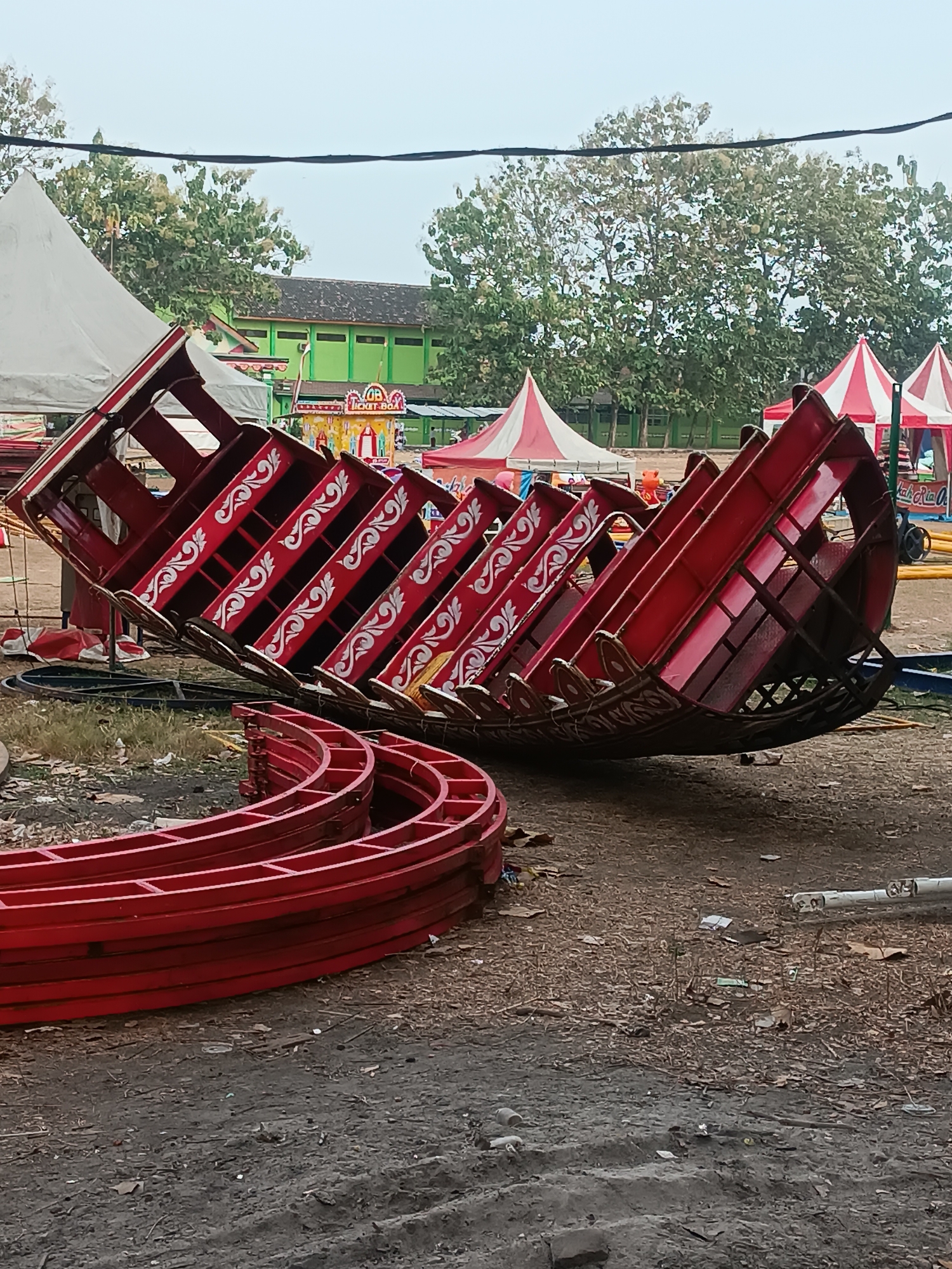 In Pleret, Indonesia, a dismantled amusement park ride is seen on the ground. The ride is red with white ornamental details on the seats. Curved red tracks are stacked in the foreground. In the background, white and red tents are set up, along with a green building. The ground is dirt and covered with fallen leaves. The sky is overcast.