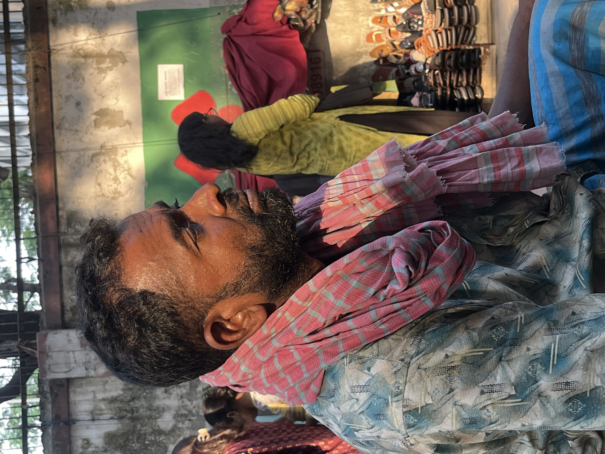 The image captures a scene in what appears to be an outdoor market area. A man with dark hair and a beard is prominently featured, wearing a patterned shirt and a red and light blue checked cloth around his neck. Several other individuals are present in the background, including a person wearing a yellow and green striped garment, another with red clothing, and additional people partially visible. Piles of sandals and shoes are stacked nearby. The setting suggests a crowded, possibly commercial environment. The location, Dhaka, Bangladesh, is based on the clothing styles and general appearance of the people.