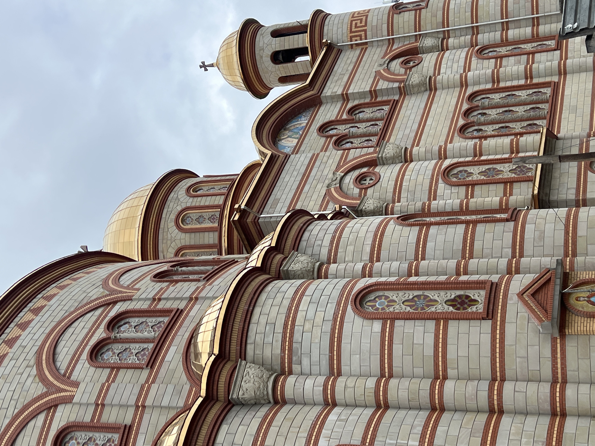 The image shows a close-up, upward-angled view of the architectural details of the Cathedral of the Immaculate Conception of the Blessed Virgin Mary in Ternopil, Ukraine. The building features a multi-tiered design with alternating bands of cream-colored and red bricks, accented by gold trim. Several domes are visible, with golden surfaces reflecting the light. The windows are decorated with stained glass or intricate floral patterns. A cross tops one of the smaller domes. The sky in the background is overcast and gray.