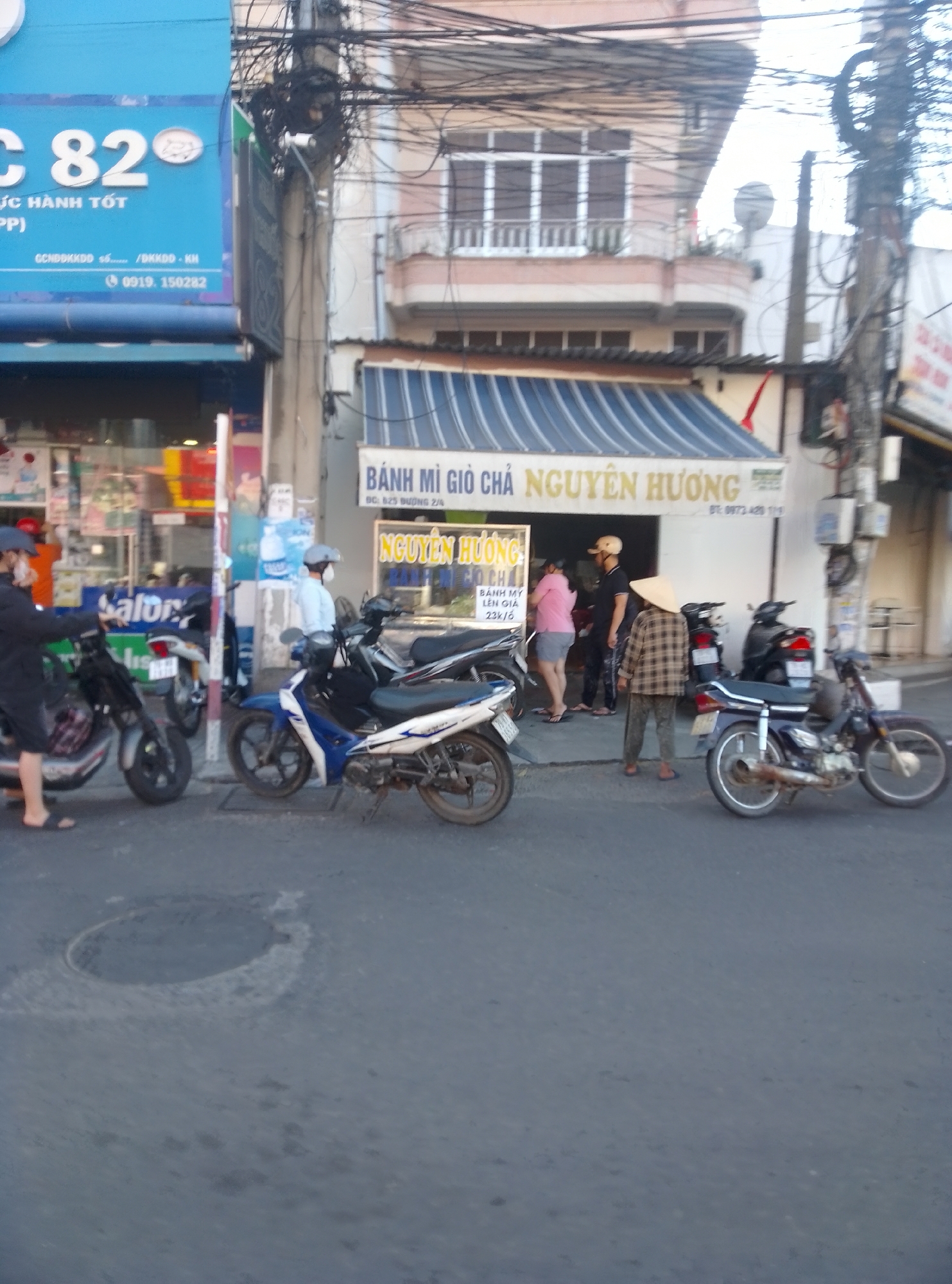 The image depicts a street scene in Bac Nha Trang Ward, Vietnam, with several people and motorbikes. In the foreground, a dark-colored motorbike is parked on the asphalt road. Further back, a person wearing a checkered shirt and a conical hat stands with a cane, facing away from the camera, near another motorbike. To the left, a person in a light blue shirt and helmet rides a white and blue motorbike. Another motorbike is parked to the left of this rider. In the background, a building with a blue awning and signage that reads "C 82" is visible. There are also other buildings and utility poles with numerous wires. Another person, wearing a pink shirt and shorts, is walking in front of a building with a sign that says "BÁNH MÌ GIÒ CHA NGUYỄN HƯƠNG." A person in a black shirt and cap stands near the entrance of this establishment. To the far left, a person in dark clothing and a mask is standing next to a motorbike. The road surface has some discoloration and a circular depression.