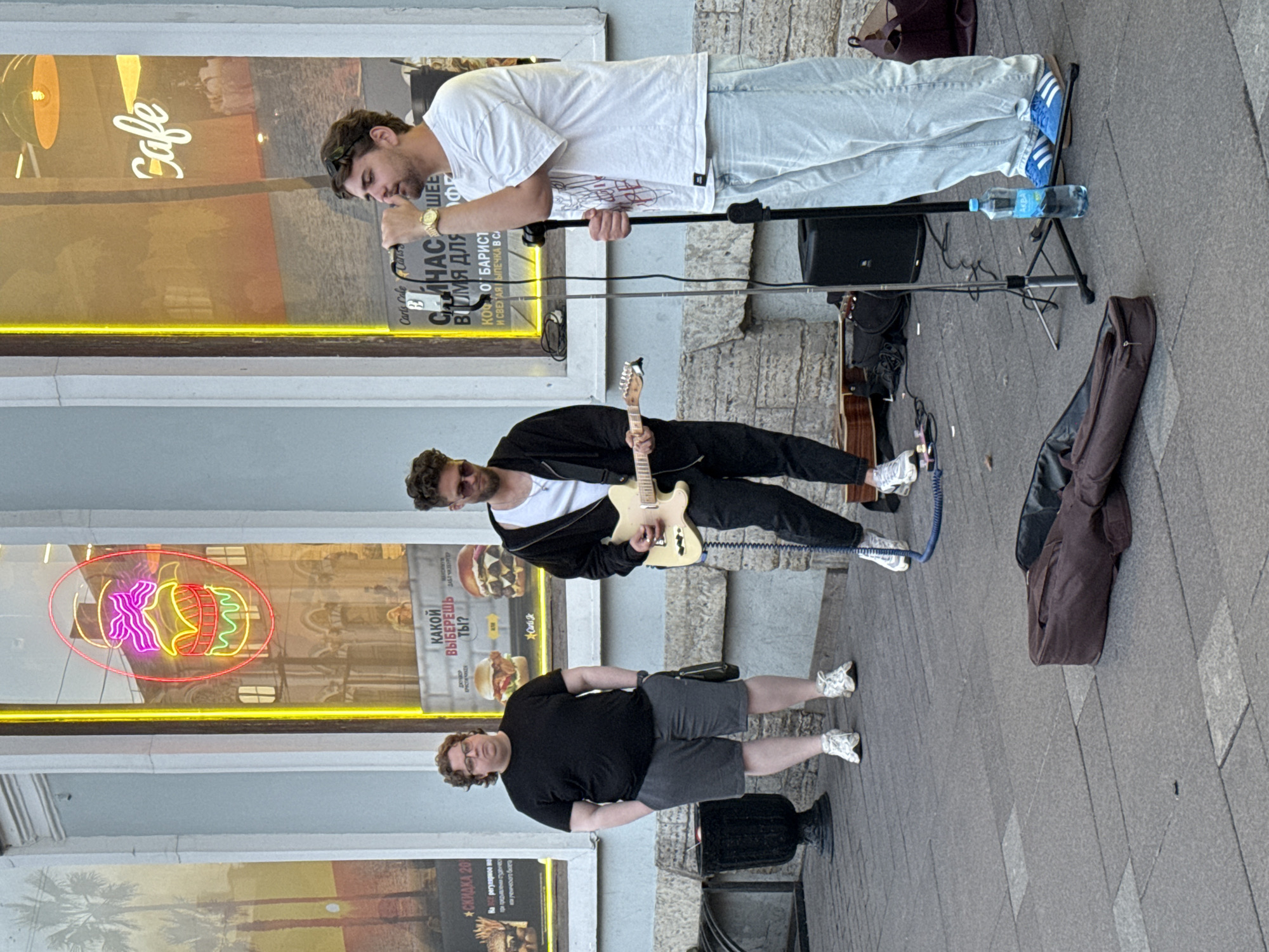 The image depicts a street scene with three men. One man in a white t-shirt and light blue jeans is standing and singing into a microphone. Another man in a black jacket and dark pants is playing a light-colored electric guitar. A third man in a black t-shirt and grey shorts is standing and appears to be observing. All three men are wearing sneakers.
In the background, there is a storefront with neon signs. One sign in red and yellow appears to be a logo with the word "Cafe" in stylized script. Another neon sign is a pink and yellow outline of food items, possibly burgers. There are also some posters on the wall, one of which displays palm trees. The ground is paved with light-colored tiles.
A black guitar case lies on the ground near the guitarist. A microphone stand with a water bottle on it is also visible. The scene appears to be taking place during the daytime, with natural light illuminating the area. The overall mood seems casual and informal, suggesting a street performance. Visible Russian text on the posters translates to "DISCOUNT 20%" and "WHAT WILL YOU CHOOSE?".