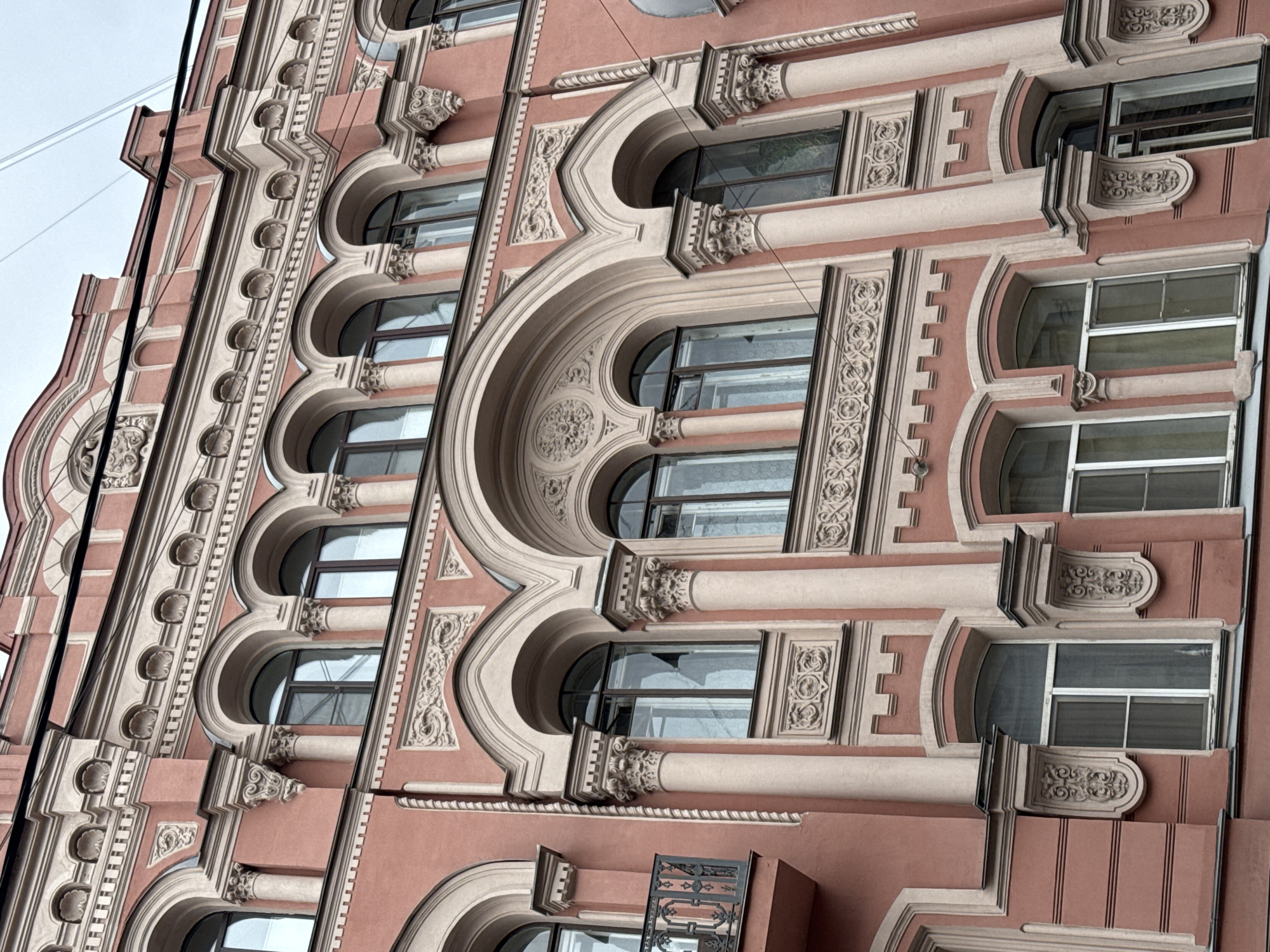 A low-angle shot reveals a building facade in Saint Petersburg, Russia. The architectural style is ornate, with a salmon-colored stucco exterior and detailed, creamy-colored moldings and window frames. The building appears to be several stories tall, with various sizes and shapes of windows, including arched and rectangular designs. Some windows have grilles or balconies visible. Elaborate decorative elements, such as carved patterns and columns, enhance the facade. Overhead wires cut across the upper left portion of the frame against a pale sky.