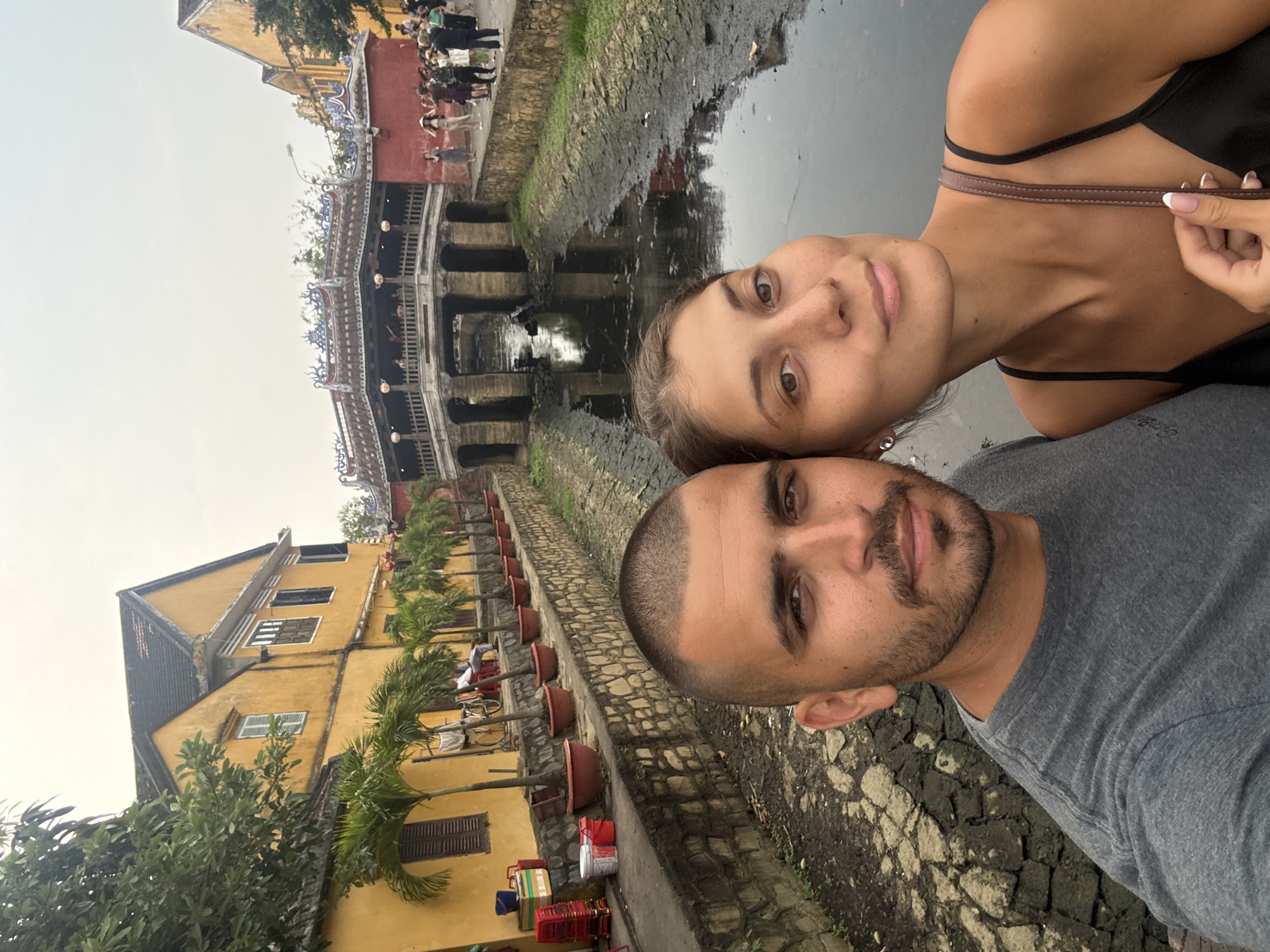A sideways-oriented selfie-style photograph captures a man and a woman in the foreground against the scenic backdrop of Hội An, Vietnam. The man, positioned on the left, has short dark hair and a beard, wearing a grey t-shirt, and looks directly at the camera with a calm expression. The woman, to his right, looks towards the camera with a subtle smile, her light brown hair pulled back, showcasing freckles and brown eyes. She is dressed in a black tank top, with her left hand lightly holding one of its straps.
Behind the two individuals, a tranquil canal dominates the mid-ground. Spanning the canal is the iconic Japanese Covered Bridge, distinguished by its traditional design, multiple arched openings, and ornate roof. Numerous people are visible on and around the bridge, indicating bustling activity. The canal banks are constructed from stone, and the water appears dark and still.
Along the left side of the canal, a row of characteristic yellow-walled buildings with dark tiled roofs stands, featuring windows and occasional balconies. A stone-paved walkway lines the canal, adorned with small palm trees planted in terracotta pots. Near the buildings, some everyday objects like crates, buckets, and red baskets are visible, hinting at local commerce.
The sky is overcast, providing soft, diffused lighting across the scene, suggesting either an early morning, late afternoon, or a generally cloudy day. The overall ambiance is one of historical charm and relaxation. No discernible text is visible within the image.
