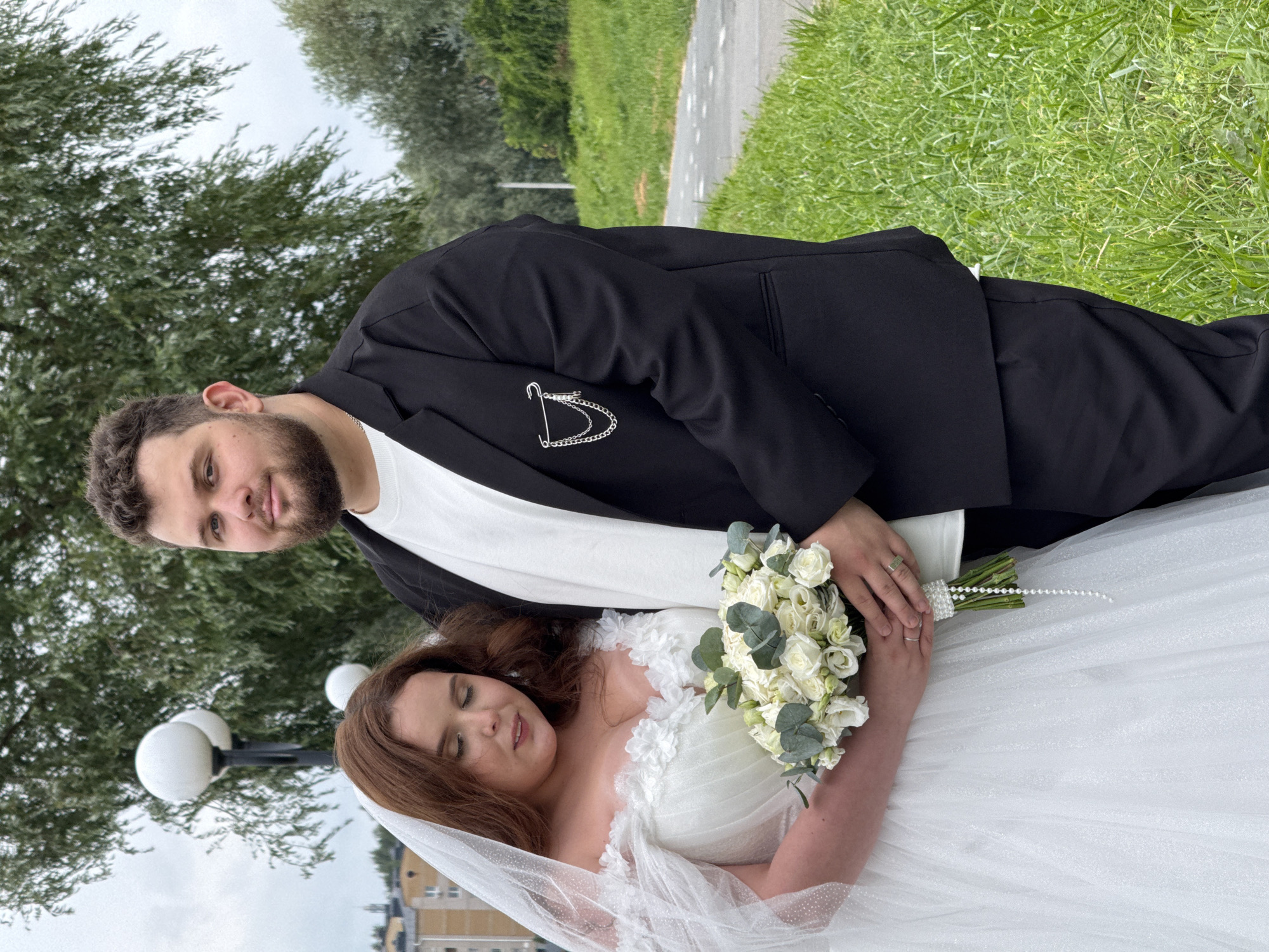 A medium shot shows a bride and groom posing outdoors in Votkinsk, Russia, on what appears to be a cloudy day. The bride, on the left, wears a white off-the-shoulder wedding dress with floral applique and a veil. She holds a bouquet of white roses and eucalyptus. The groom, on the right, is dressed in a black suit jacket over a white t-shirt and dark pants. He has a chain detail pinned to his lapel. They are standing on a grassy area next to a paved path. In the background, trees and a distant building are visible.