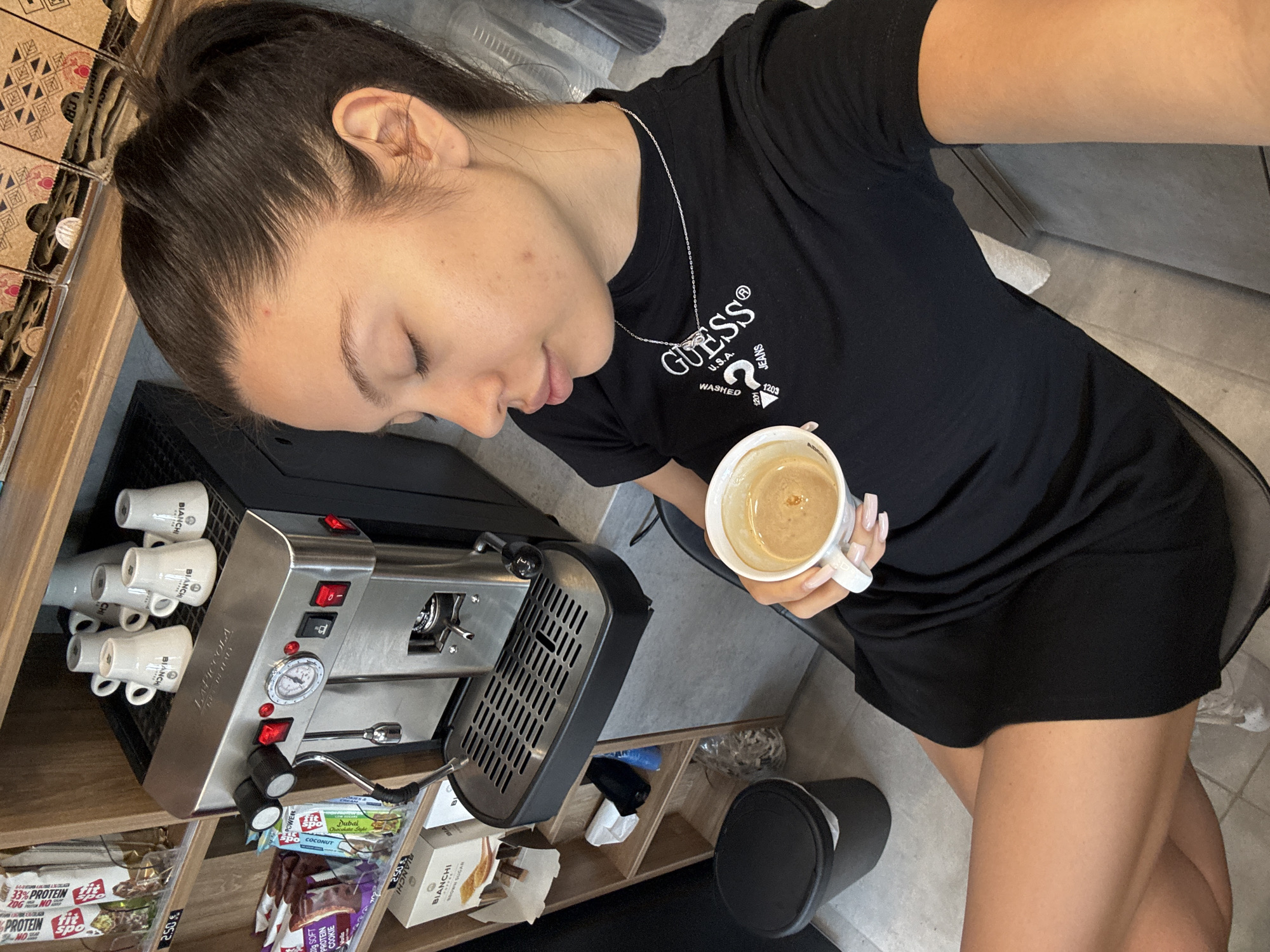 A young woman with dark hair pulled back in a ponytail sits in a chair, holding a cup of coffee. She is wearing a black t-shirt with a white "Guess" logo and a silver necklace. The coffee cup has a white handle and appears to be full of coffee. The woman is looking down at the coffee, with her eyes closed.
In the background, there is a coffee machine with several cups on top of it. There are also shelves with various items, including what appear to be protein bars and boxes of coffee or tea. A black trash can is visible to the left of the coffee machine. The environment appears to be an indoor cafe or shop.
The scene is consistent with a cafe in Sofia, Bulgaria.