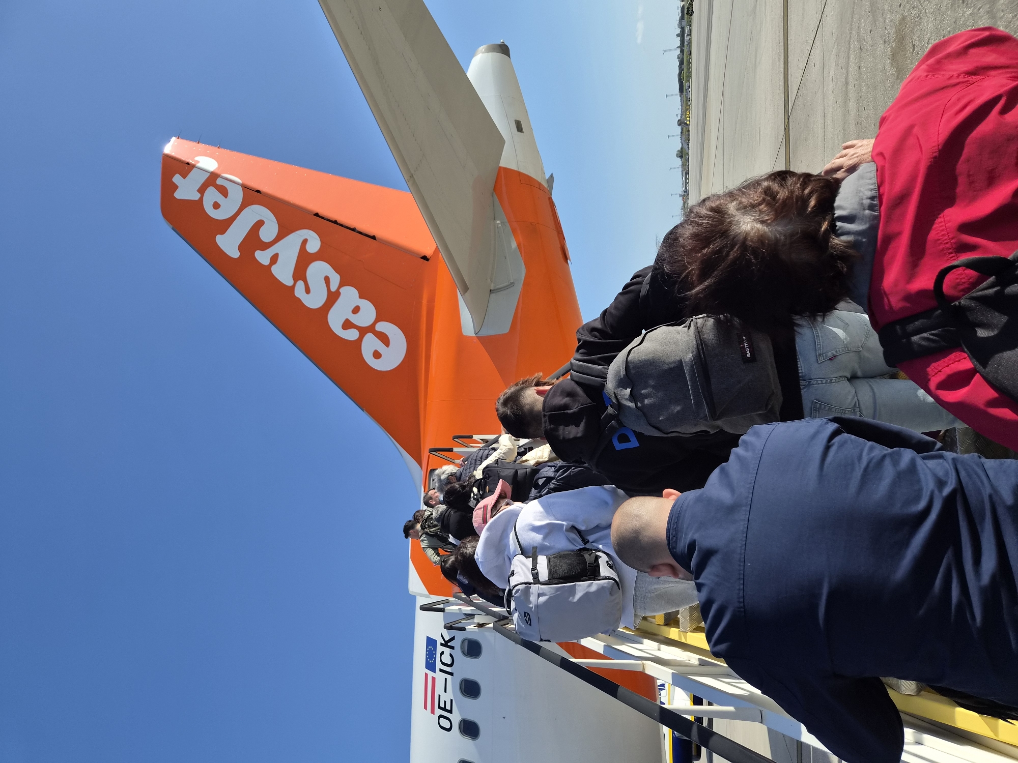 Passengers are boarding an EasyJet airplane, evidenced by the tail fin displaying the company logo. The aircraft is marked with "OE-ICK," indicating its registration. Passengers are ascending the stairs towards the plane's entrance. The location may be the city of Moreira, Portugal, but this is impossible to ascertain from the image alone. The day is clear with a blue sky. The apron is visible.