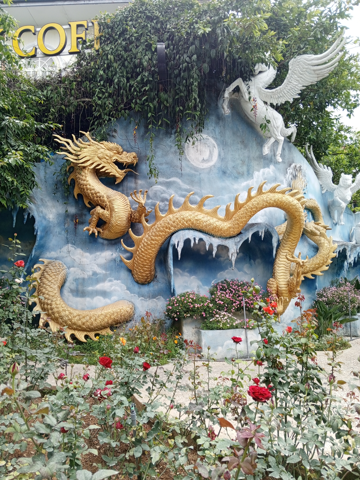 The image shows a decorative wall sculpture in Da Lat, Vietnam, featuring a large golden dragon coiled around an icy landscape. Behind the dragon, a white Pegasus with outstretched wings is mounted on the wall. The wall is painted with a sky and cloud motif, with a large moon visible. The sculpture is situated in an outdoor setting with a garden of red and pink roses and other flowers in the foreground. Vines and foliage hang from the top of the wall, partially obscuring a sign that reads "COFF". The scene is set against a cloudy sky.