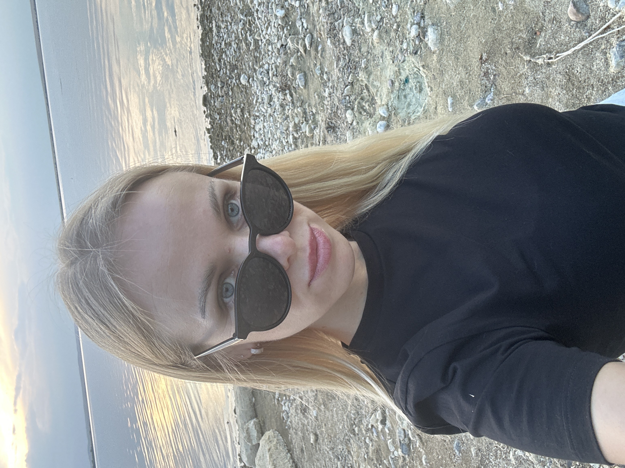A blonde woman wearing black sunglasses and a black t-shirt poses for a selfie at a rocky shoreline. The sun is setting over a body of water, casting a warm glow on the clouds and water. The woman's hair is long and blonde, and she has blue eyes. She is wearing hoop earrings with small stones. The background is a mix of water, rocks, and sand.
