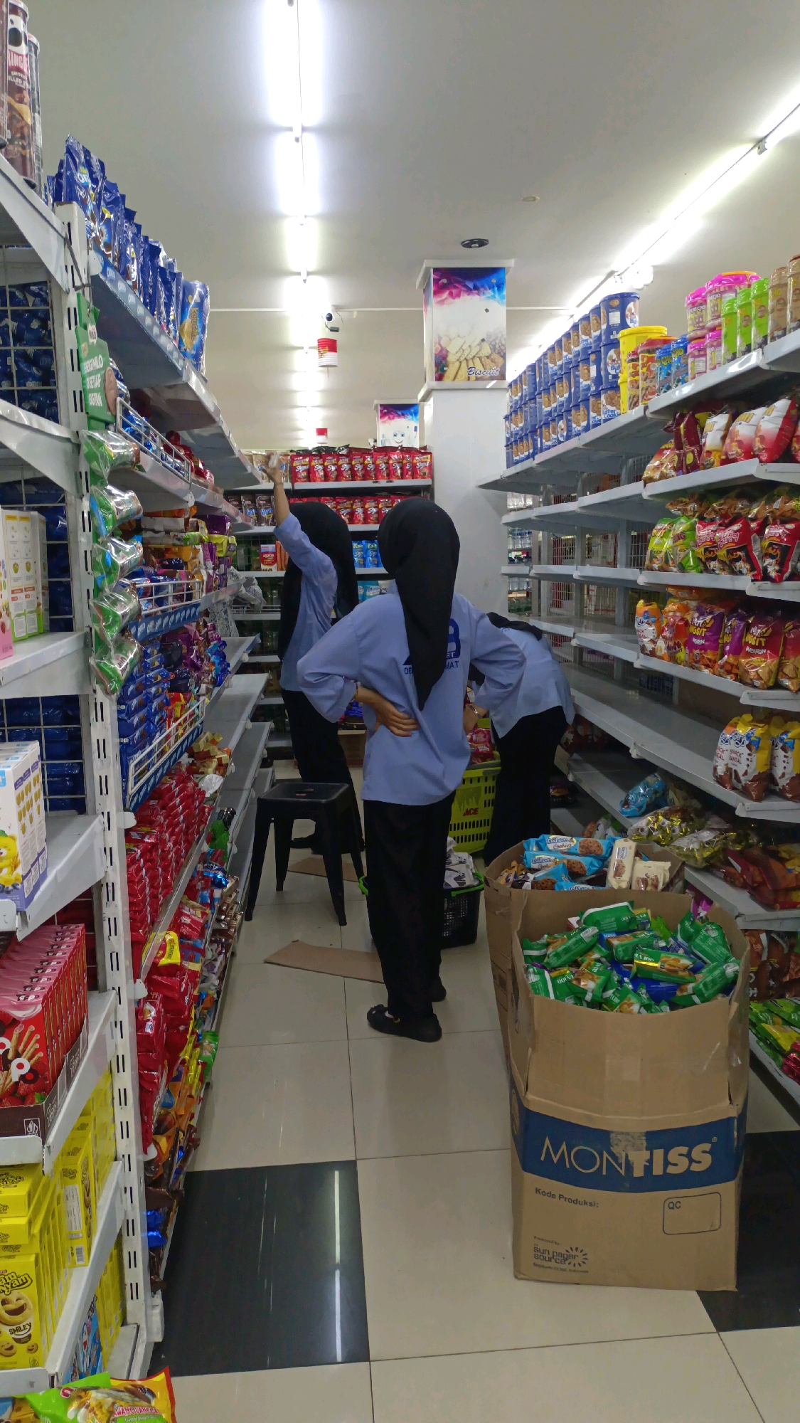 The image depicts the interior of a convenience store in Padang Panjang, Indonesia. Three individuals, presumably employees, are visible. One individual, wearing a light blue shirt and black headscarf, is standing with their back to the camera, hands on hips, facing a shelf stocked with food items. Another individual, also in a light blue shirt and black headscarf, is seen from the side, reaching up towards a higher shelf. The third individual, similarly attired, is partially visible in the background, bent over. The store is well-stocked with merchandise arranged on shelves lining the aisles. Prominent displays include packaged snacks, biscuits, and drinks. A large cardboard box, labeled "MONTISS," filled with green-packaged items, is placed on the floor in the foreground. The flooring is tiled in a checkered pattern of black and light beige squares. Overhead lighting illuminates the scene.