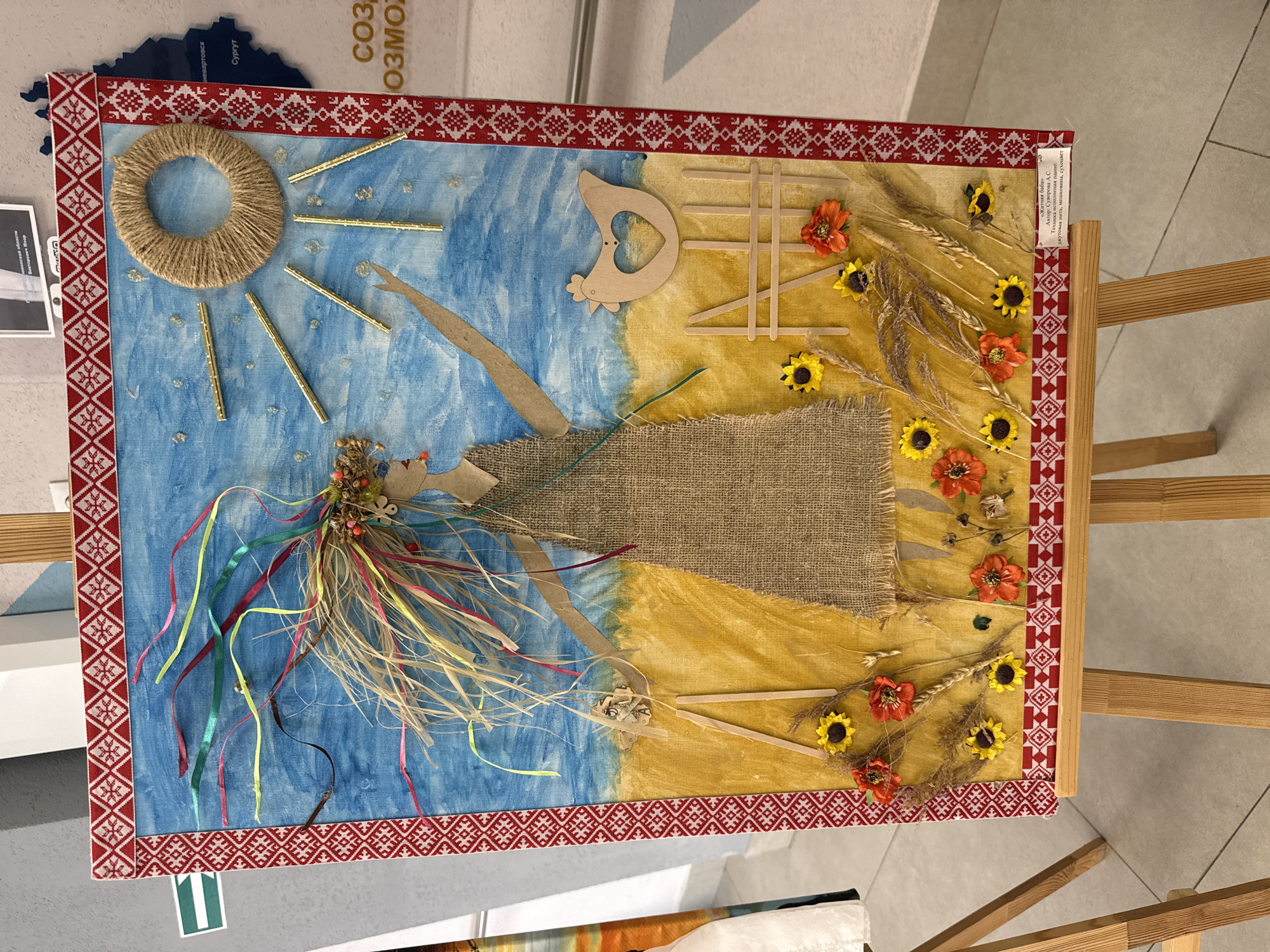 The image shows an artwork titled "Rye Woman" by A.S. Suvorova from Surgut, Russia. The artwork is a mixed-media piece, utilizing materials such as jute thread, burlap, and dried flowers. It depicts a figure on a background divided into blue and yellow sections, possibly representing the sky and a field. The figure, constructed with burlap for the torso and sticks for the arms and legs, has a headdress of dried flowers and ribbons. A jute ring and golden sticks represent the sun in the upper left. A wooden chicken shape and wooden sticks are placed on the right side of the panel. The entire artwork is bordered with a red and white patterned trim. The artwork is displayed on a wooden easel. A label on the right side of the panel identifies the artist and materials used. The artwork is located in Tobolsk, Russia, according to the text on the top left corner of the image.