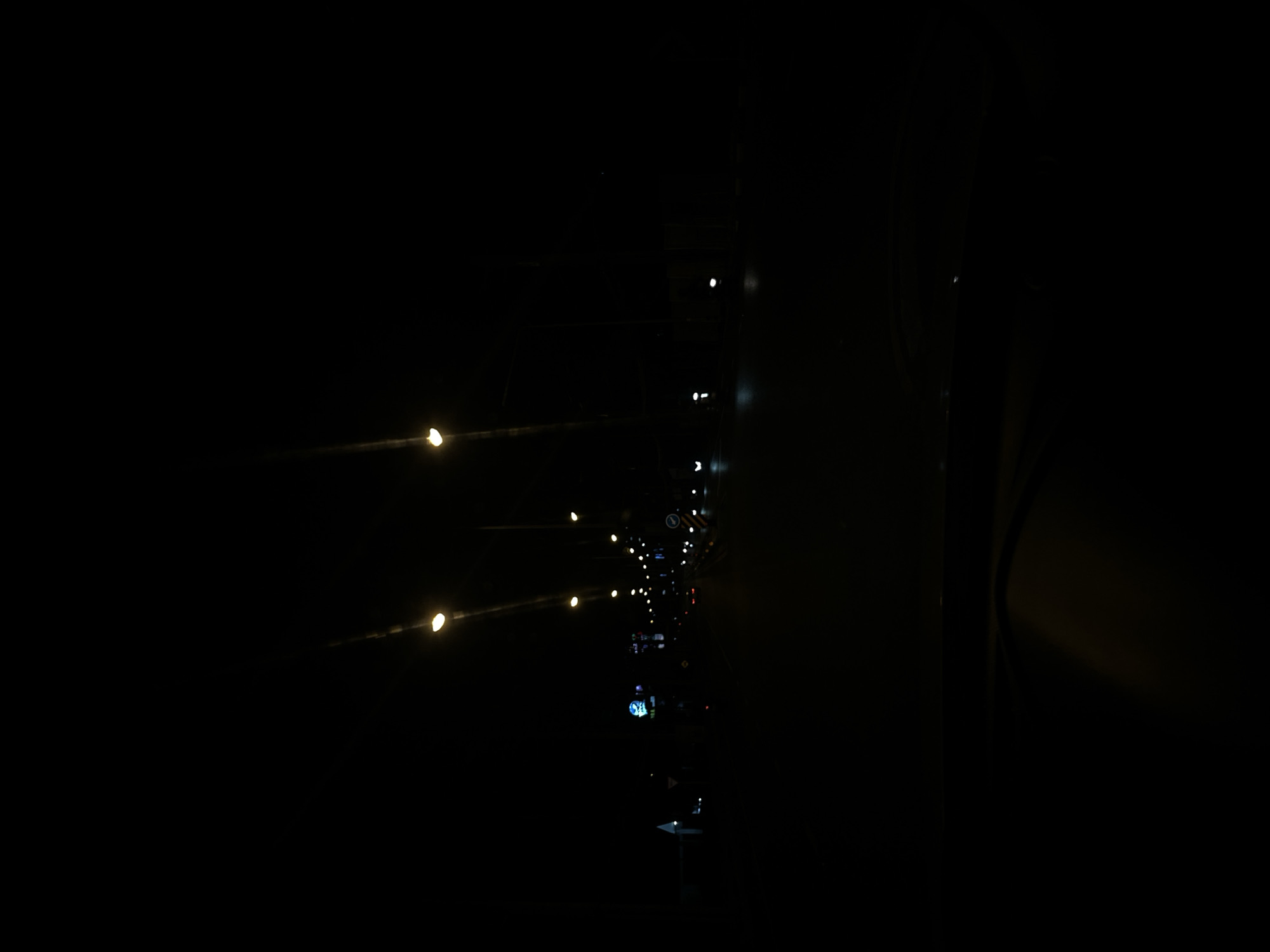 This is a night-time image taken outdoors in Chalong, Thailand. The scene is very dark, with several illuminated objects scattered throughout. On the left side of the frame, there are several bright, blurry lights that appear to be streetlights or vehicle lights, stretching diagonally from the bottom left upwards and to the right. In the center and right side of the image, there is a vertical arrangement of smaller lights and some faint structures. These lights are more distinct and appear to be a mixture of white and possibly some colored lights, suggesting traffic signals, illuminated signs, or building lights. There are also some visible signs with text, including what looks like a circular road sign with yellow and black stripes at the top right, and a blue sign with white text further down. The overall impression is of a dimly lit urban or semi-urban environment at night, possibly near a road or intersection. The darkness and the nature of the lights suggest a low light condition, possibly with some atmospheric haze. No people are visible in the image.