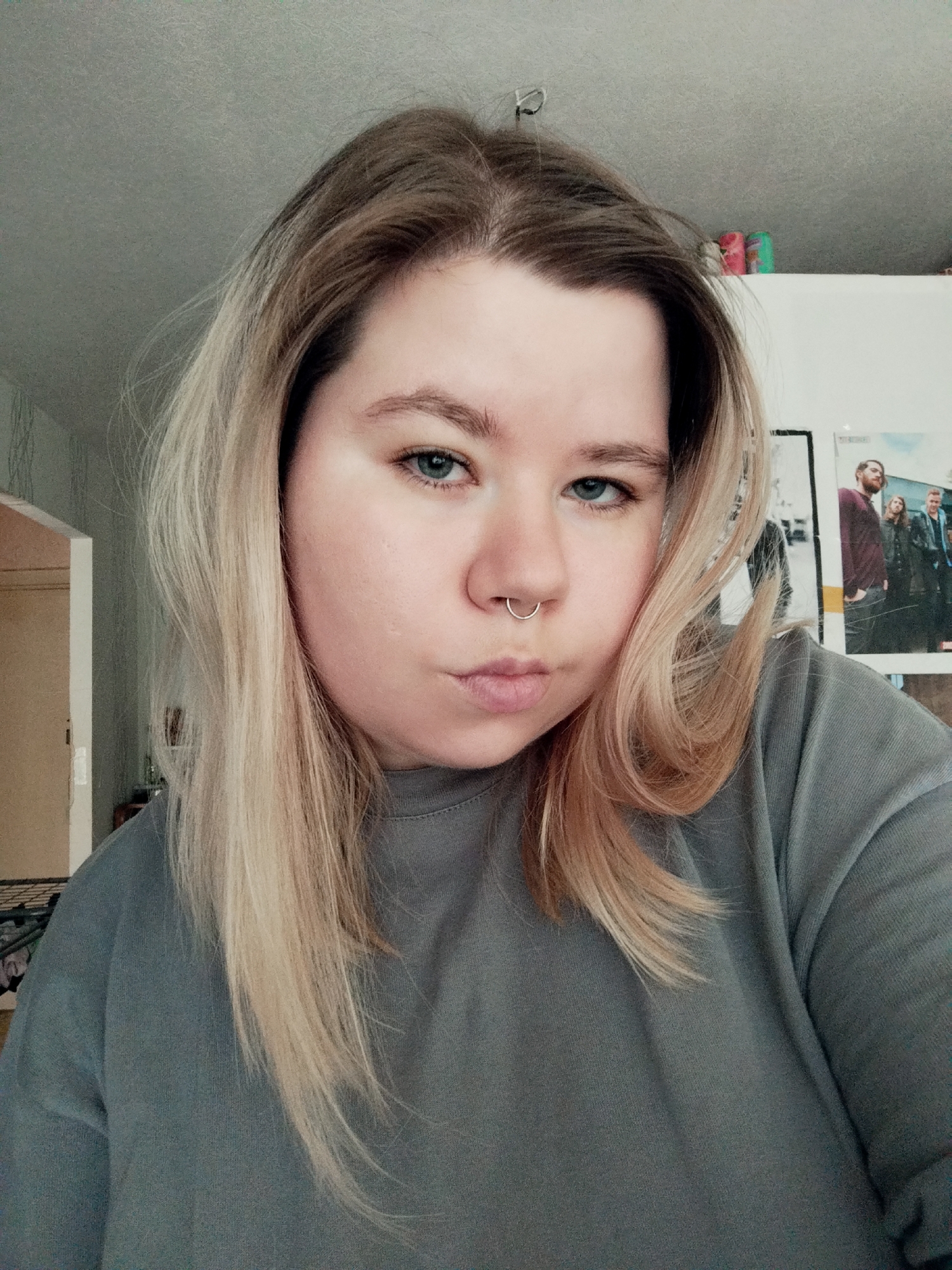 A selfie captures a woman with fair skin, light brown hair with blonde highlights, and blue eyes. She has a septum piercing and is wearing a gray sweatshirt. Her expression is neutral, with slightly pursed lips. The background includes a white wall with posters and items on a shelf above. To the left, a doorway is visible, leading to another room. The location is Unknown, Unknown.