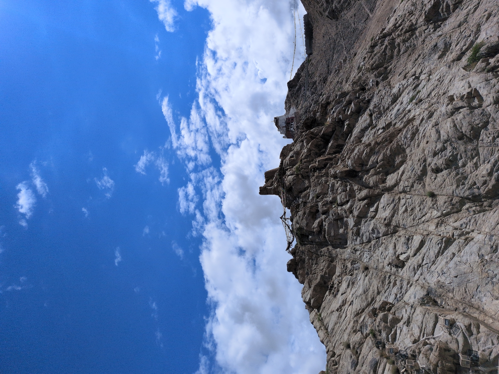 This image captures the stark beauty of the Leh region in India, likely showcasing a prominent landmark like the Leh Palace or a similar monastery perched atop a rocky outcrop. The foreground is dominated by a rugged, rocky hillside, its surface textured with countless cracks and crevices, suggesting a natural geological formation. Sparse patches of vegetation cling to the rock face. A series of small, square enclosures, possibly part of a prayer or marker system, are visible ascending the left side of the hill.
Further up the incline, a weathered structure, likely an ancient monastery or palace, stands defiantly against the elements. It features whitewashed walls with darker trim and a prominent, multi-tiered architecture, indicative of traditional Himalayan building styles. Prayer flags are visible strung between some of the rocky peaks near the structures, their colorful fabric fluttering in the breeze. In the distance, another section of a fortified wall or walkway can be seen connecting to another elevated point.
The sky above is a vibrant, deep blue, dotted with fluffy white clouds. The lighting suggests a bright, sunny day, casting sharp shadows on the rocky terrain and highlighting the textures of the landscape and the architecture. The overall impression is one of ancient grandeur set against a dramatic, arid mountain environment. No people are visible in the frame.