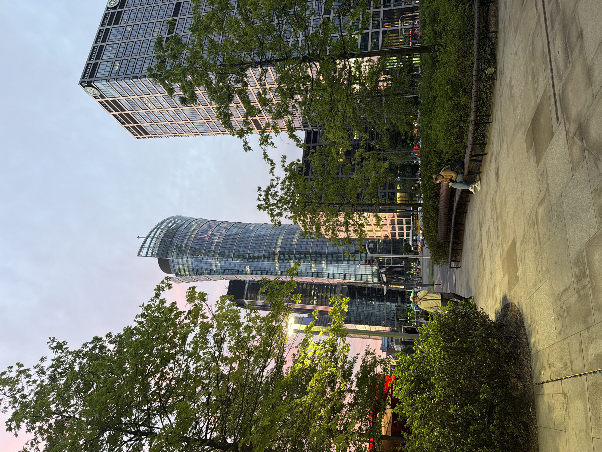 The image shows an urban environment in Unknown, Unknown, featuring modern architecture and greenery. Two high-rise buildings with glass facades dominate the skyline. One building has a curved top, and the other is a rectangular grid. In the foreground, trees with green foliage frame the buildings. Along a paved walkway, two people are sitting on a bench, and another person is standing near a hedge. The sky is a blend of pink and grey, suggesting either sunrise or sunset. Streetlights provide illumination.