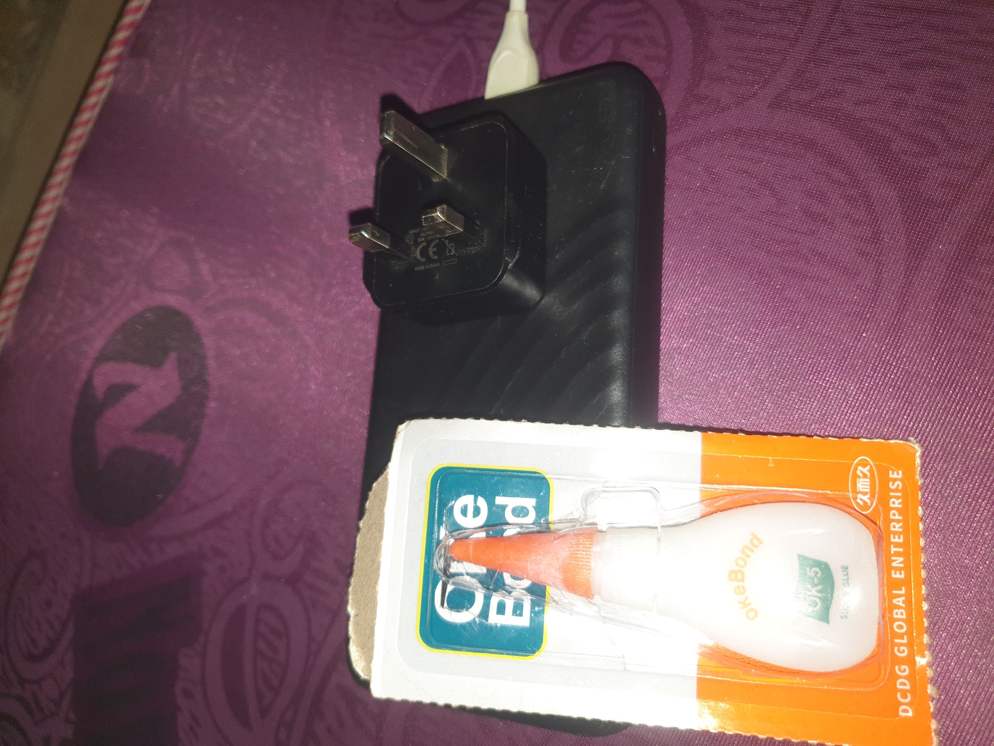 In a close-up, top-down shot, a black power bank is positioned on a textured purple surface. A white USB cable is plugged into the power bank. Resting on top of the power bank is a black electrical adapter with three silver prongs, characteristic of a UK plug. To the left of the power bank, a blister pack containing a small bottle of "One Bond" super glue is visible. The glue bottle has an orange tip and white body with text that includes "OK-5" and "SUPER GLUE." The background of the power bank and the surface beneath it are a deep purple with a subtle pattern. The overall scene is domestic or travel-related, suggesting a charging setup with an adhesive product nearby.