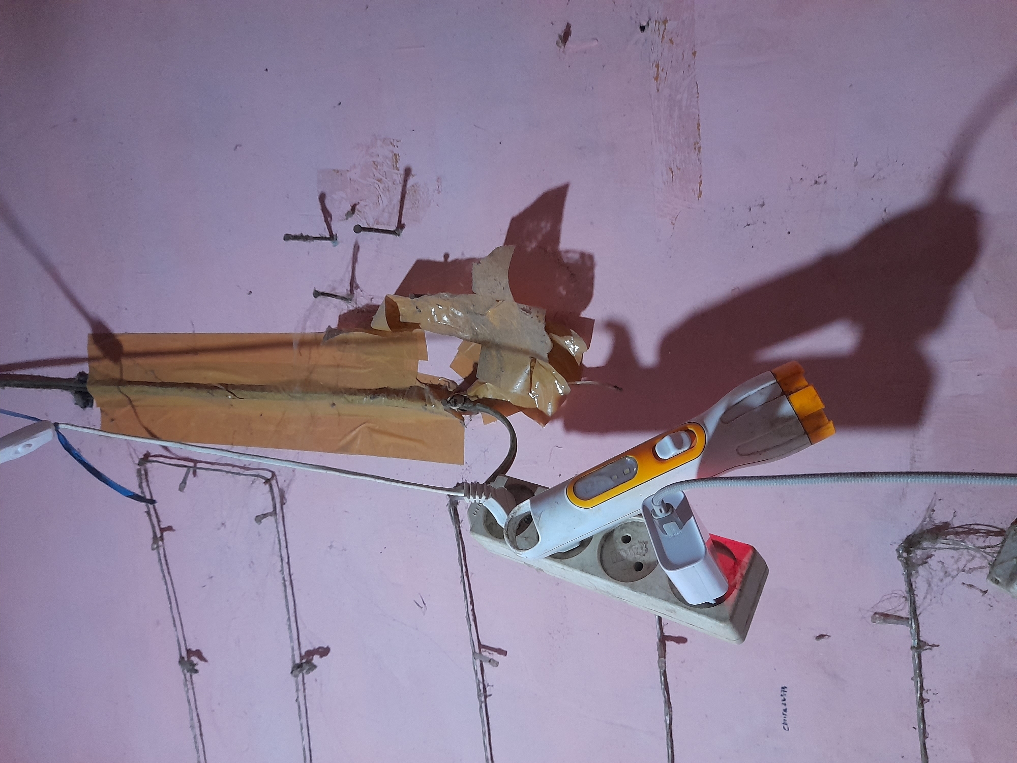 The media file shows a close-up, slightly angled view of a pink wall with visible damage and repair attempts. A long, beige electrical extension cord with multiple outlets is mounted horizontally on the wall. A white and orange flashlight, with its LED lights illuminated, is plugged into one of the outlets. A white adapter is plugged into another outlet. Wires are visible along the wall, some secured with nails and others appearing loose. A piece of orange tape is applied to the wall above the extension cord, covering some of the damage and a cable. Another piece of torn, tan tape is also visible. The lighting suggests an indoor setting, possibly dimly lit, with a shadow of the flashlight and possibly a person's head and shoulders cast on the wall. The wall appears rough and textured, with peeling paint and what look like remnants of older plaster. Small nails are hammered into the wall in several places, likely to guide or secure wires. The overall impression is one of an unfinished or deteriorating electrical setup. No specific time of day or weather conditions are discernible. The text "CHINA LUSA" is faintly visible on the wall near the bottom right of the frame. The location context is Pulokulon, Indonesia.