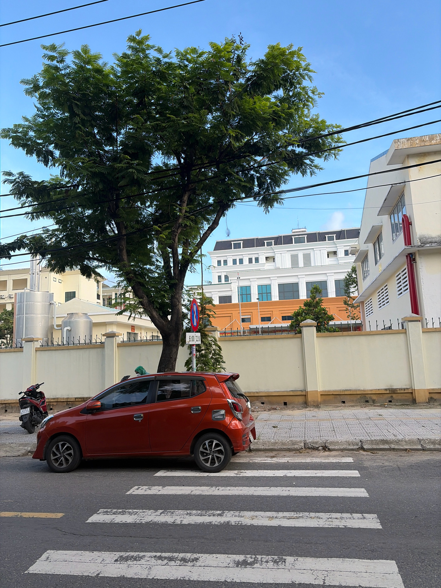 The image captures a street scene in Da Nang, Vietnam. A bright orange compact car is parked on the right side of the road, partially on a pedestrian crosswalk. To the left of the car, a red motorcycle is parked along the sidewalk. Behind the sidewalk, a cream-colored wall with a black metal fence runs horizontally across the frame. A large tree with lush green foliage stands in the middle ground, casting shadows on the car and sidewalk. Beyond the wall and tree, several buildings are visible. One building, directly behind the tree, appears to be a multi-story structure with a white facade and dark-tiled roofs. To the right of this building, another white building with red accents is partially visible. In the background, a larger white building with multiple windows and a darker roofline dominates the skyline. The sky above is a clear blue with some thin clouds. A traffic sign with red and blue markings and a time indicator (6:00-22:00) is visible on a pole near the tree. The street surface is asphalt, marked with white pedestrian crossing stripes. The overall impression is a typical urban street environment in Vietnam.