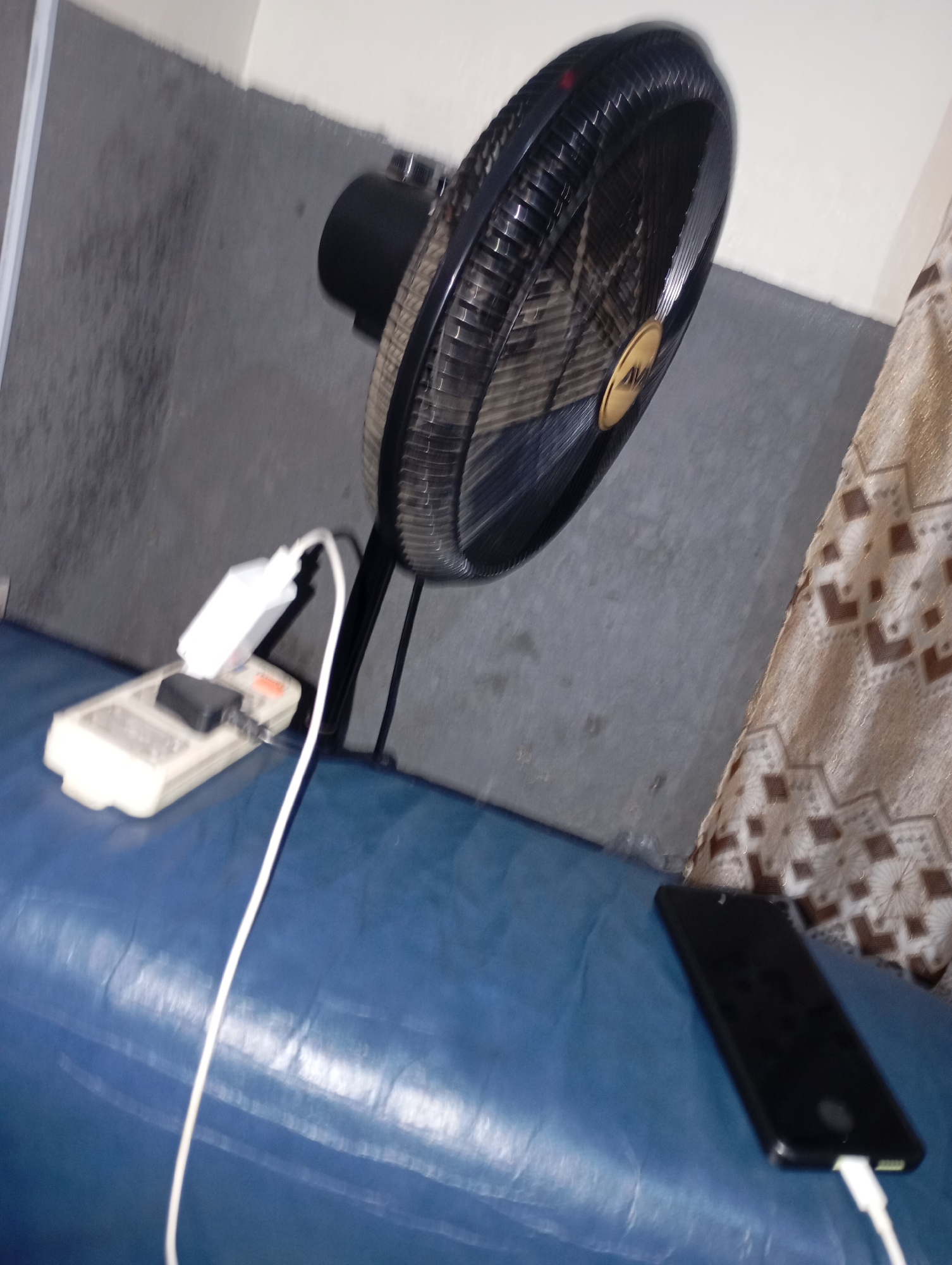 The image depicts a close-up, somewhat blurry view of a desk or table surface in an indoor setting. A black electric fan with a gold emblem is positioned on the upper right side, angled downwards. To the left of the fan, a power strip is visible, with a white electrical adapter plugged into it, and a black object also connected. A white electrical cord extends from the adapter and runs across the surface. Further to the right, a black smartphone with a white charging cable plugged into its bottom is lying on the surface. The background consists of a textured gray wall. A patterned fabric is visible on the far right, draped behind the fan and smartphone. The surface itself appears to be a blue, possibly vinyl, material. The lighting is dim, contributing to the overall blurriness.