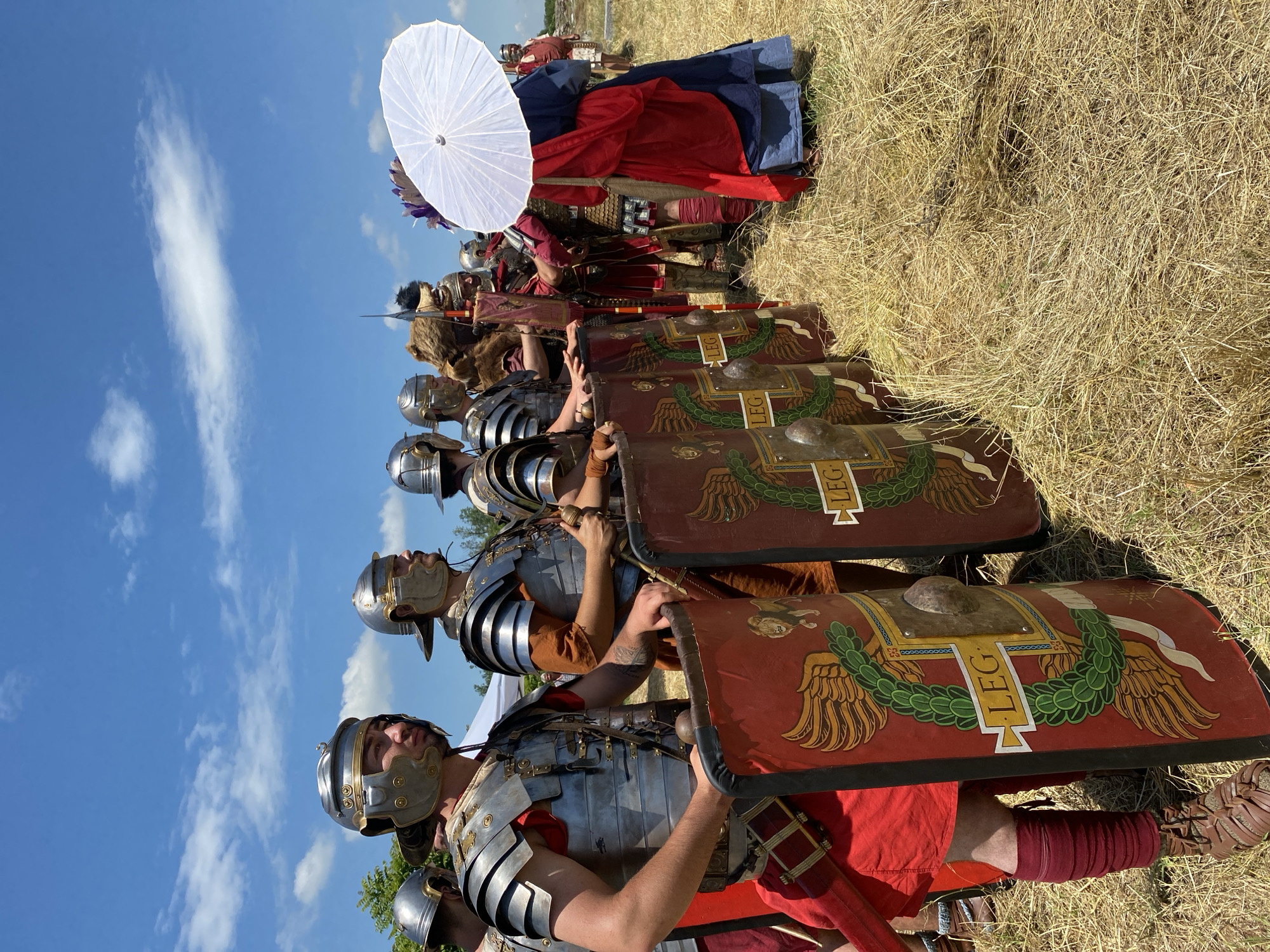 The image depicts an outdoor historical re-enactment scene under a clear blue sky with scattered white clouds, likely occurring in Gigen, Bulgaria, possibly at a site associated with the Roman castrum Oescus.
In the foreground and mid-ground, a line of individuals dressed as Roman soldiers, or legionaries, stands on dry, golden-brown straw or hay-covered ground. The lead figure on the left is a male wearing a silver-colored segmented plate armor (lorica segmentata) over a red tunic, a Galea helmet with brass accents, red leg wraps, and brown leather caligae (sandals). He holds a large, rectangular red scutum (shield) upright with his left hand, displaying a central metal boss, a golden winged design, and a green laurel wreath encompassing a white banner inscribed with "LEG." A visible tattoo is present on his right forearm.
To his right, additional legionaries are similarly attired, each holding an identical red scutum with the "LEG" inscription. One individual in the middle ground wears a bear pelt over his armor, holding a vertical staff or standard. Further to the right, a person in a blue garment with a red draped sash and a multicolored feathered or plumed headpiece is partially obscured by a large, white circular parasol held by another individual.
In the background, additional figures in various historical costumes are visible, along with green foliage from trees or bushes on the left and a white tent or canopy. The scene is brightly lit by natural daylight, indicating a sunny outdoor event.