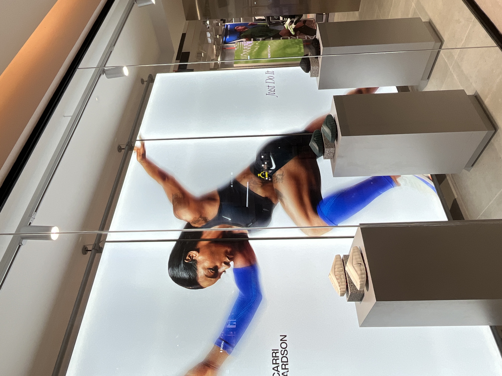 The image shows a Nike store window display. The main subject is a large illuminated advertisement featuring a female athlete in motion, likely a runner. She is wearing athletic apparel, including blue arm sleeves and knee-high socks. Text on the advertisement includes "CARRI" and "Just Do It."
In front of the advertisement, on display pedestals, are several pairs of Nike shoes. The shoes are minimalist in design and appear to be a light beige color, with one pair also featuring a dark green color.
The store environment is visible through the window, with more displays and shelving in the background, as well as a green banner. The lighting appears to be artificial, with spotlights directed at the display. The floor is tiled.
There are no people present in the immediate foreground or interacting with the display. The scene is clean and organized, typical of a retail environment.
The image does not contain any information indicating its location in Semarang, Indonesia.