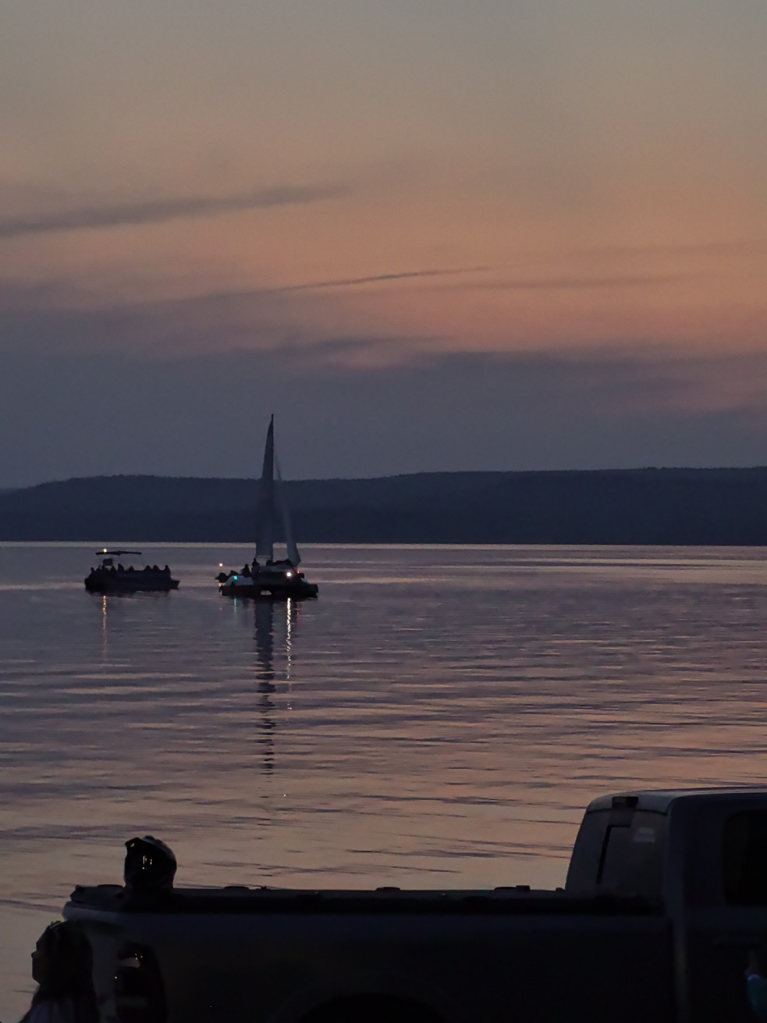 The image depicts a serene twilight scene on a body of water, likely a lake or bay, as indicated by the calm surface and distant, silhouetted shoreline. The sky displays a gradient of soft colors, transitioning from a pale orange and pink near the horizon to a muted purple and blue overhead, suggesting a sunset or sunrise.
In the mid-ground, two vessels are visible on the water. To the left, a smaller, darker boat appears to be carrying multiple passengers, their silhouettes visible against the water's reflection. To the right, a sailboat with its mast and sail silhouetted against the sky is also carrying passengers. Both boats are illuminated by lights, casting reflections on the water.
In the foreground, the silhouette of a pickup truck is prominent. The truck's bed is visible, and a dark object, possibly a helmet or a piece of equipment, rests on it. To the left of the truck, the upper body and head of a person are visible, looking towards the water. Their attire is dark, blending with the shadows. The water in the foreground exhibits gentle ripples, reflecting the colors of the sky.
The overall impression is one of quiet activity on the water during twilight, with the truck in the foreground suggesting a vantage point or a place of rest by the shore. The absence of specific Osa, Russia, landmarks in the image makes it impossible to pinpoint the exact location within the city. However, the elements present are consistent with a waterfront area.