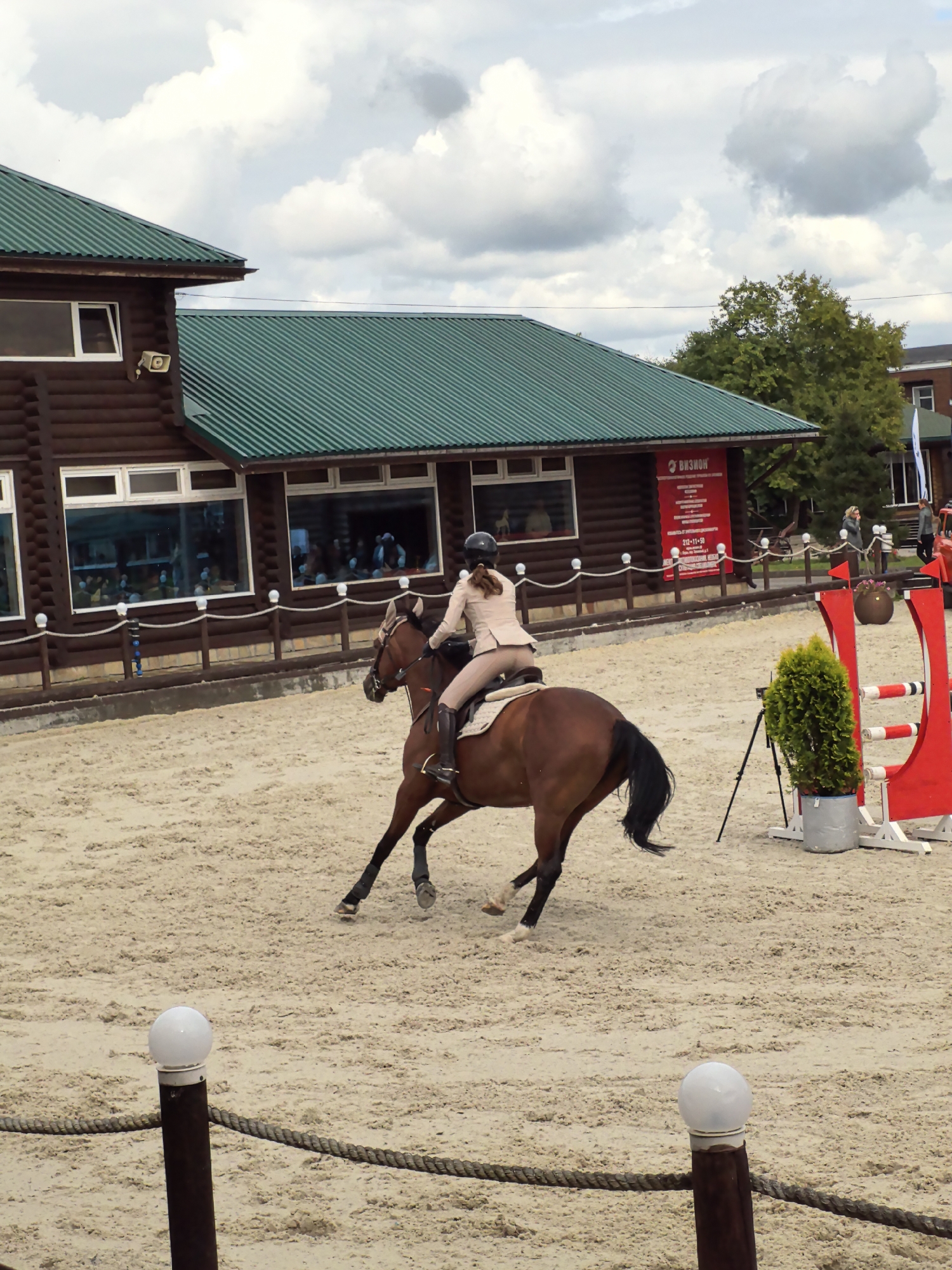 An outdoor equestrian event is underway at a farm in the city of Ferme, Russia. A person is riding a brown horse on a sandy arena. The rider is wearing a tan riding suit and helmet. The horse is in mid-stride, with its legs extended. In the background, a wooden building with a green roof is visible. The building has several large windows, through which spectators can be seen. There is also a red jump in the foreground. The sky is cloudy.